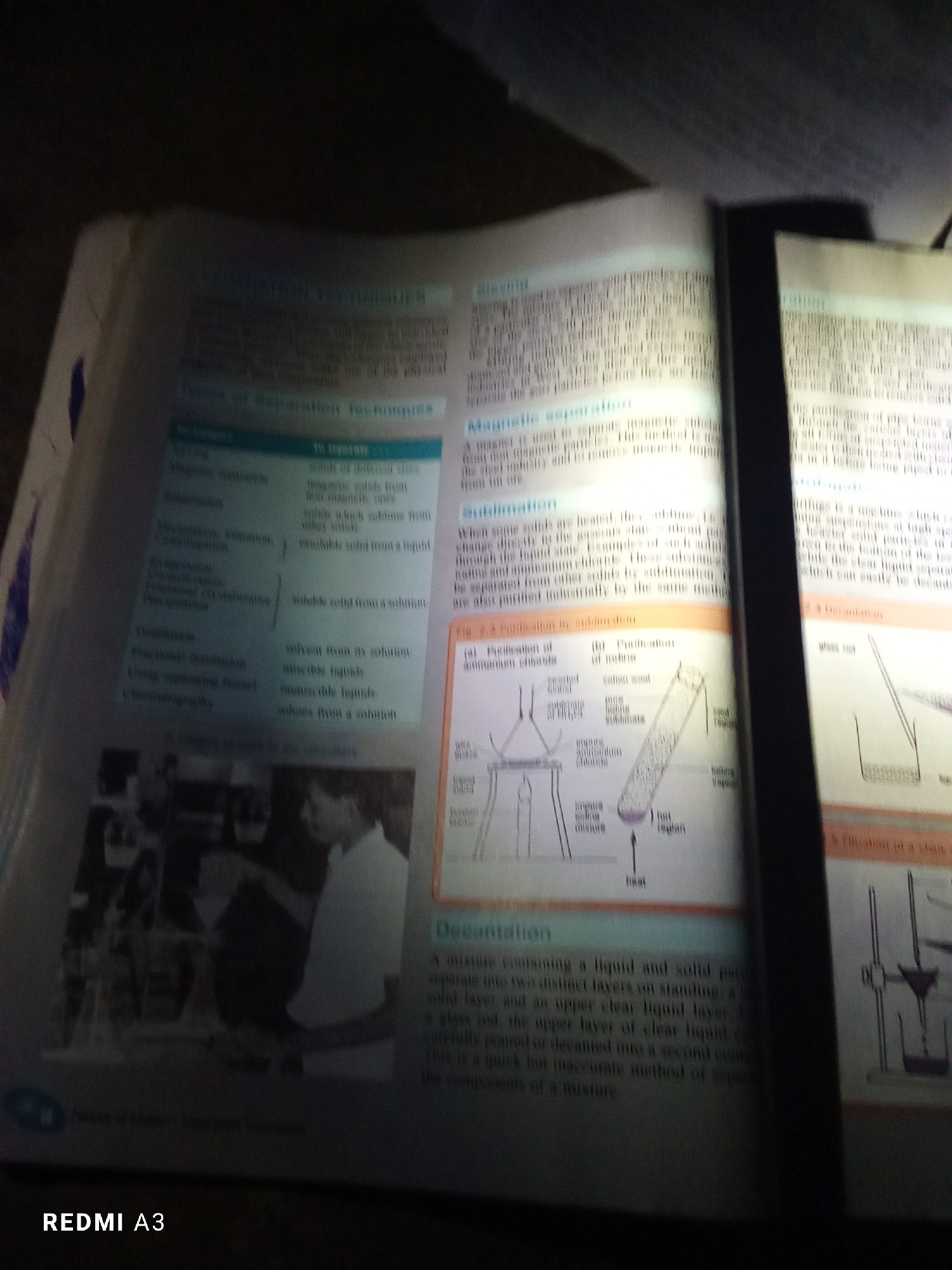 The image shows an open book with text and diagrams related to separation techniques. The book is illuminated by a light source, creating shadows and glare. The text discusses methods like sieving, magnetic separation, sublimation, crystallization, evaporation, distillation, and chromatography. Diagrams illustrate processes such as purification by sublimation and filtration. A black object, likely a phone or other electronic device, is positioned to the right of the book, obscuring part of the pages and casting a shadow. The bottom left corner of the image displays the text "REDMI A3". There is no information within the image that indicates the location in the city of Jalingo, Nigeria.