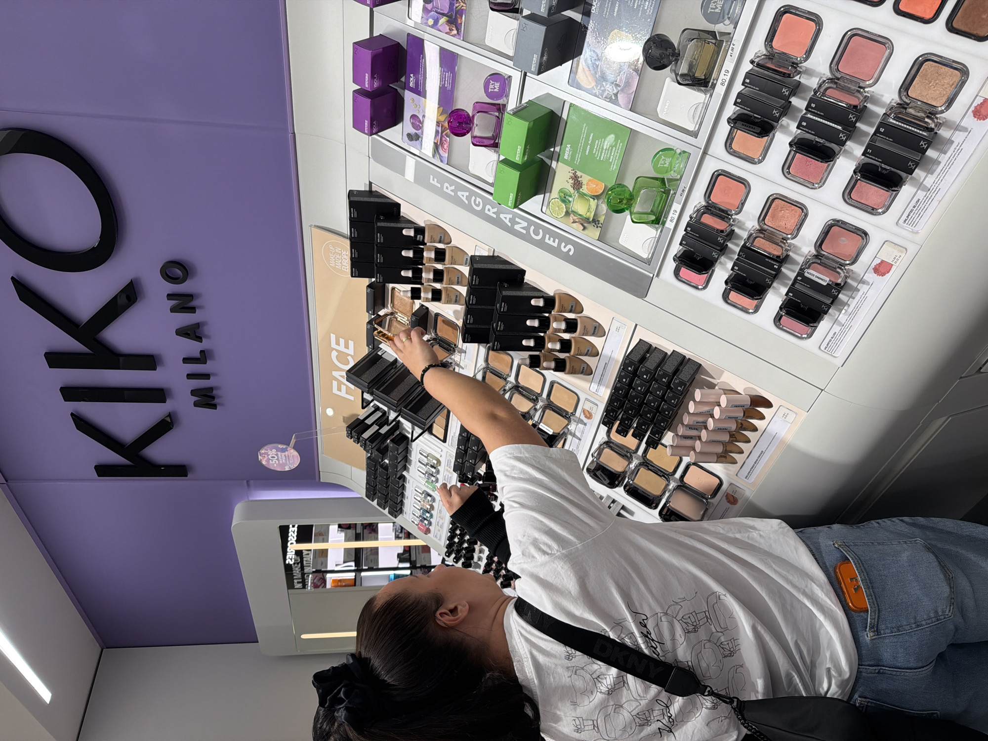 The image shows the interior of a KIKO Milano store in Burgas, Bulgaria. A woman is standing in front of a makeup display, examining the products. The display features various makeup items, including blush, foundation, and fragrance samples. The store's branding is prominently displayed, with the KIKO Milano logo on a purple wall. The woman is wearing a white t-shirt with a design, blue jeans, and a black purse. The lighting is bright and artificial, typical of a retail environment. The scene focuses on the customer's interaction with the store's merchandise.