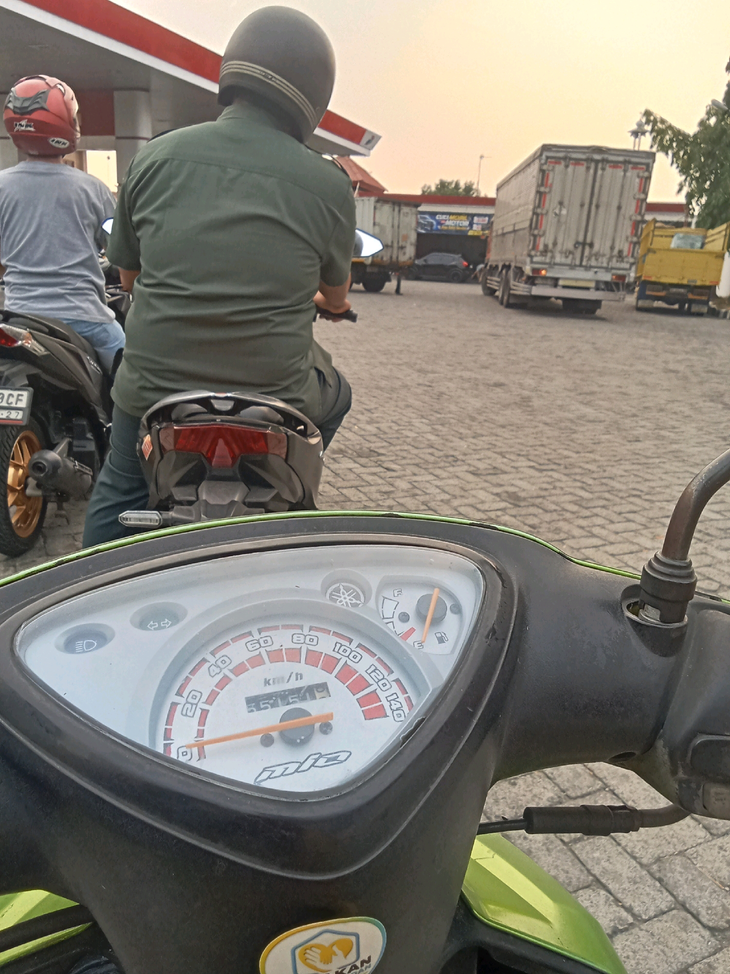 This is a first-person perspective, medium shot image captured at a gas station in Jelakombo, Indonesia. The foreground is dominated by the dashboard and handlebars of a green scooter, with a speedometer showing 55509 km/h as the current speed. The speedometer dial is marked with speed increments from 0 to 140 km/h. The scooter's handlebars are black, and a rearview mirror is attached to the right side.
In the mid-ground, two individuals are seated on a black motorcycle, facing away from the camera. The rider in the back is wearing a dark green uniform and a dark helmet. The person in front is wearing a light grey t-shirt and a red and black helmet. Their motorcycle's license plate is partially visible and reads "BCF 27." The motorcycle is positioned on a cobblestone-like paved surface, typical of outdoor areas in Indonesia.
The background features a gas station canopy with red and white stripes, and several large trucks. One truck is a white semi-trailer truck, and another appears to be a yellow dump truck. The sky is a pale orange, indicating it might be sunrise or sunset. The overall environment suggests a busy transportation hub.