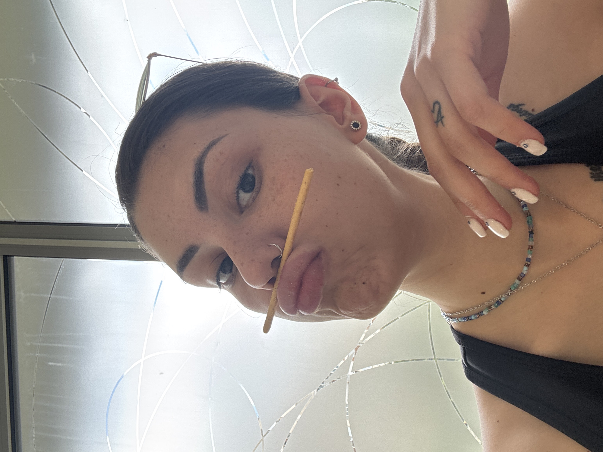 The image is a rotated close-up selfie of a young woman, taken from a low angle looking upwards. She is positioned diagonally across the frame, looking directly at the viewer with a playful, slightly pouty expression. A thin, light-brown stick, resembling a breadstick or similar snack, is held comically between her upper lip and nose.
The woman has dark hair, pulled back, with glasses perched on her head. Her face is visible with distinct freckles, and she has a nose ring. An earring with a dark center and a lighter, possibly floral, border is visible in her right ear. She is wearing a black top, possibly a swimsuit or tank top. Around her neck, she wears two layered necklaces: one a delicate, multi-colored beaded choker in blues, greens, and earthy tones, and another a simple silver chain. Her fingernails are painted a pale color, likely white or light pink, and feature small decorative dots. A small, stylized tattoo, which appears to be a symbol like a Libra sign, is visible on her finger. A faint, indistinct tattoo is also partially visible on her upper torso.
The background consists of a bright, diffused light source coming from what appears to be a large window or skylight. The glass features distinctive white, curved, and linear patterns, suggesting either frosted glass or a decorative overlay. The bright, natural illumination indicates it is daytime. The overall mood of the image is casual and humorous. No explicit text is visible beyond the stylized finger tattoo. The location context is Sofia, Bulgaria, though no specific landmarks are visible in the image itself.