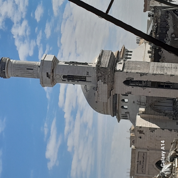 The image displays the heavily damaged Al-Farouq Mosque in Khan Yunis, Palestinian Territory, under a blue sky with scattered white clouds. The mosque's light-colored minaret, topped with a fluted cylindrical section, shows significant structural damage on its lower rectangular base, with visible crumbling masonry and exposed internal structures. The mosque's main dome, also light-colored, exhibits numerous pockmarks and surface damage. Adjacent to the minaret and dome, the mosque's main building is severely compromised, with large sections of walls collapsed or missing, revealing arched openings and extensive rubble at ground level. An inscription in Arabic script is visible on a surviving wall segment to the left of the dome. The immediate foreground shows a chaotic pile of debris and rubble, suggesting widespread destruction. A dark, diagonal structural element, possibly a damaged beam or rebar, occupies the right foreground. No individuals are present in the scene. The image bears a "Galaxy A14" watermark in the bottom-left corner.