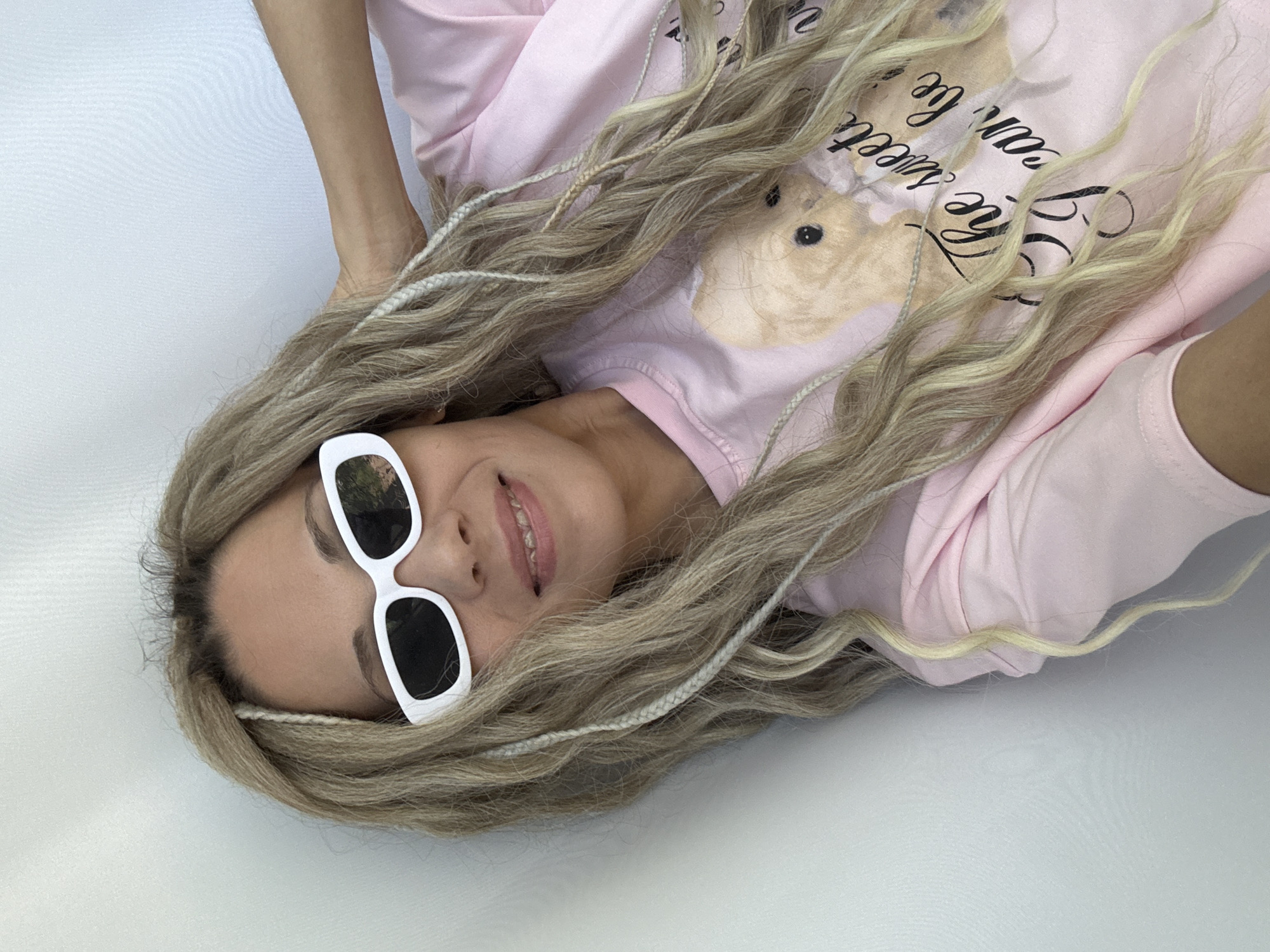 A woman with long, wavy blonde hair with white braids interspersed lies on a white surface. She is wearing white-framed sunglasses with dark lenses. Her hair partially covers a light pink long-sleeved shirt with a graphic of a hamster and the words "The Sweetest One" printed on it. The location is not discernible from the image.