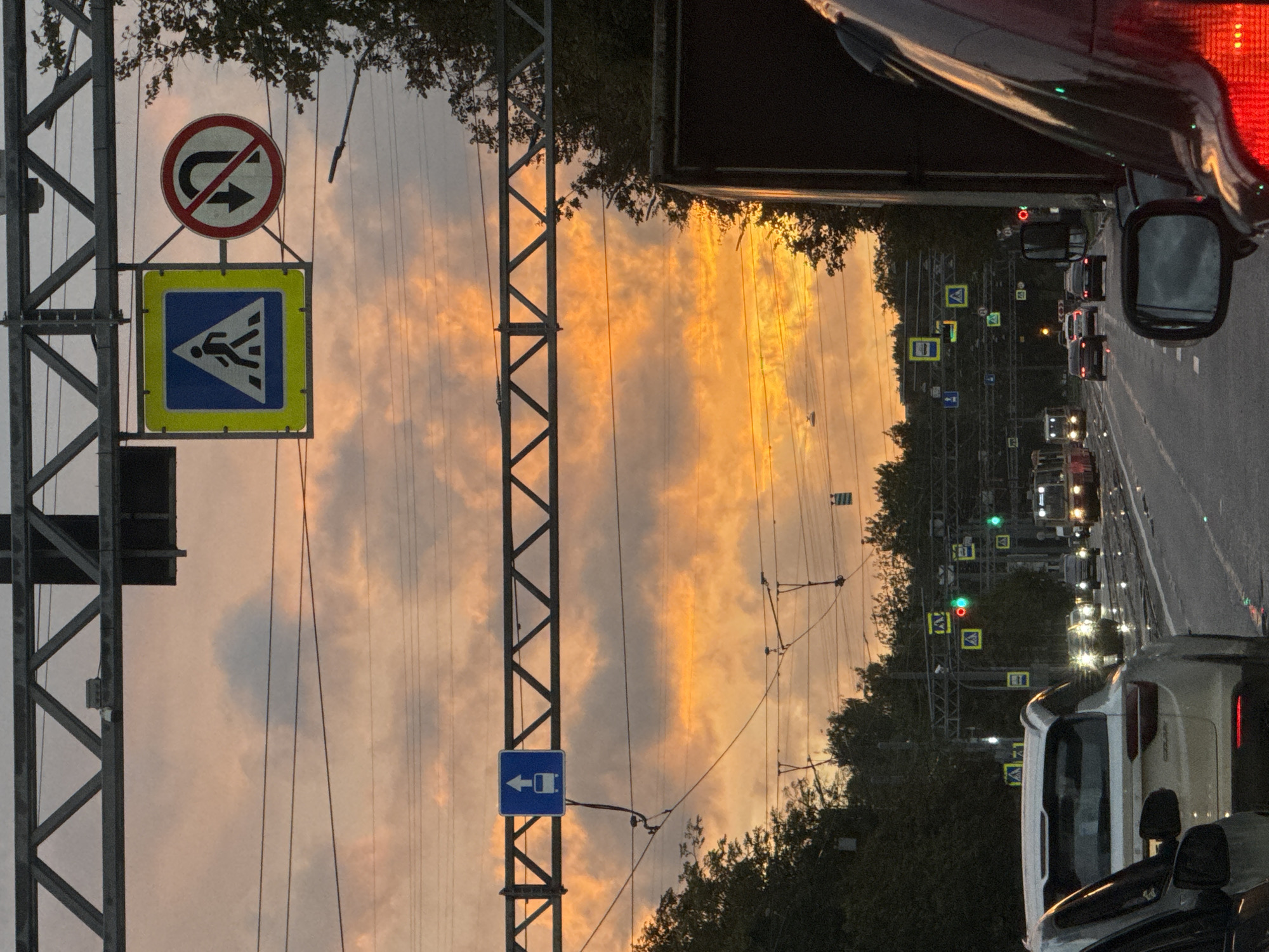 The image depicts a street scene at dusk in Sinyavino, Russia, with a dramatic sky filled with orange and pink clouds. The foreground features the rear of a white SUV and the side mirror of a dark-colored vehicle. A traffic-filled road stretches into the distance, with a tram visible on the left and several cars. Overhead, a metal truss structure supports various traffic signs, including a pedestrian crossing sign, a no U-turn sign, and a sign indicating a bus lane. Trees line the sides of the road, and power lines crisscross the sky. The overall impression is that of a busy urban environment at the end of the day, with a striking natural backdrop.