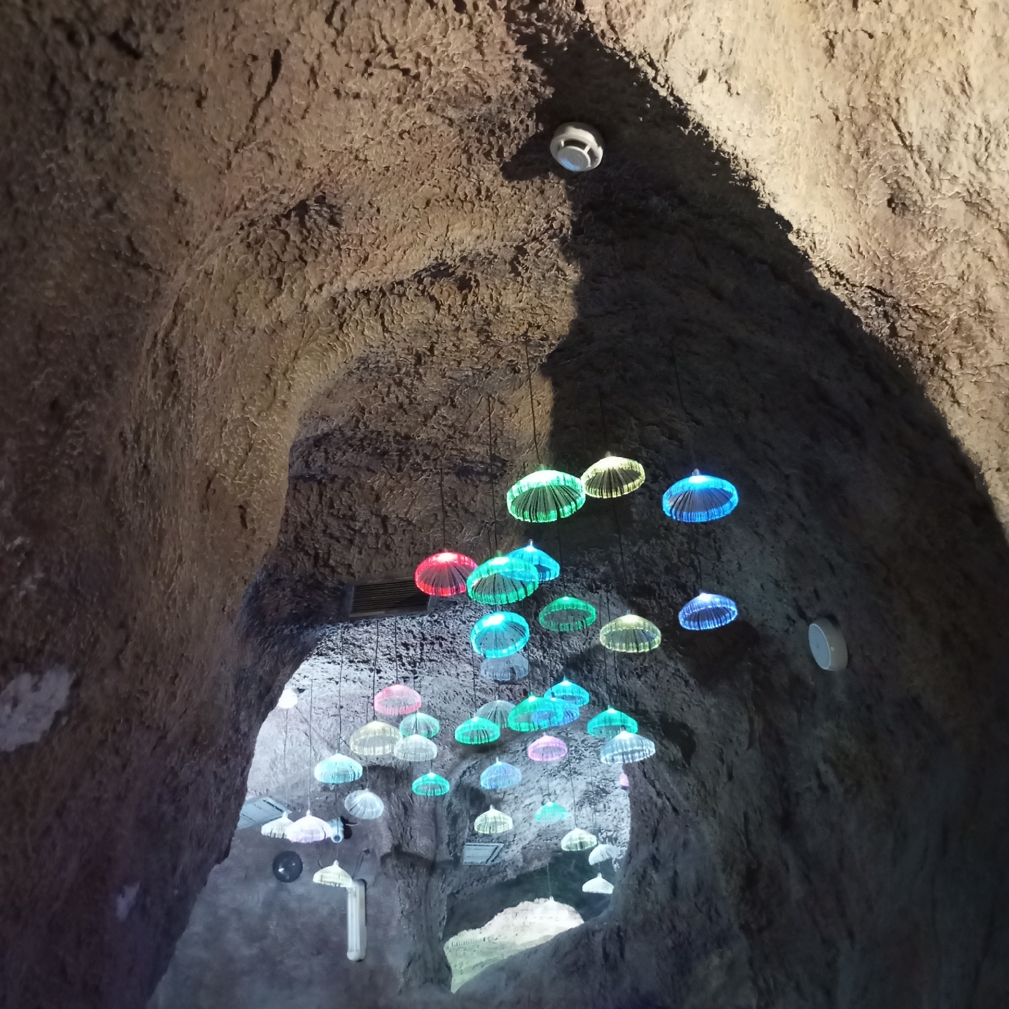 The image depicts an indoor cave-like environment with textured, rough walls that are dimly lit. Hanging from the ceiling by thin wires are numerous decorative light fixtures. These fixtures are shaped like translucent jellyfish or umbrellas and are illuminated with various colors, including green, blue, red, and pink, creating a vibrant and whimsical atmosphere. In the upper right portion of the image, a white smoke detector is visible mounted on the wall. A white circular speaker is also visible on the right wall. The overall impression is that of an art installation or themed area within a building. This scene is located within the "Skypark" attraction in the city of Sochi, Russia, a popular adventure park situated in the Akhshtyr Gorge.