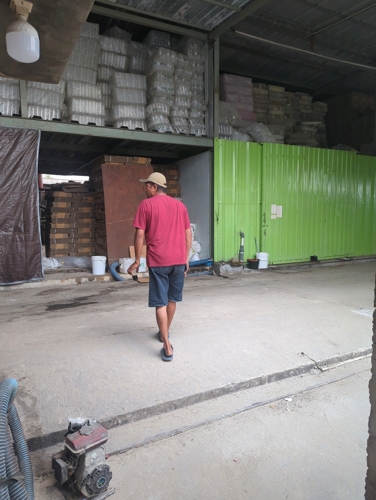 A person wearing a red t-shirt, blue shorts, and a tan baseball cap walks away from the camera in a warehouse. The warehouse has stacked goods on shelves, including what appear to be plastic containers and boxes. A large green metal wall is visible on the right side of the image, with pipes and buckets near its base. In the foreground, a blue hose and a small engine-powered device are on the concrete floor. The scene is located in Batu Belaman, Indonesia.