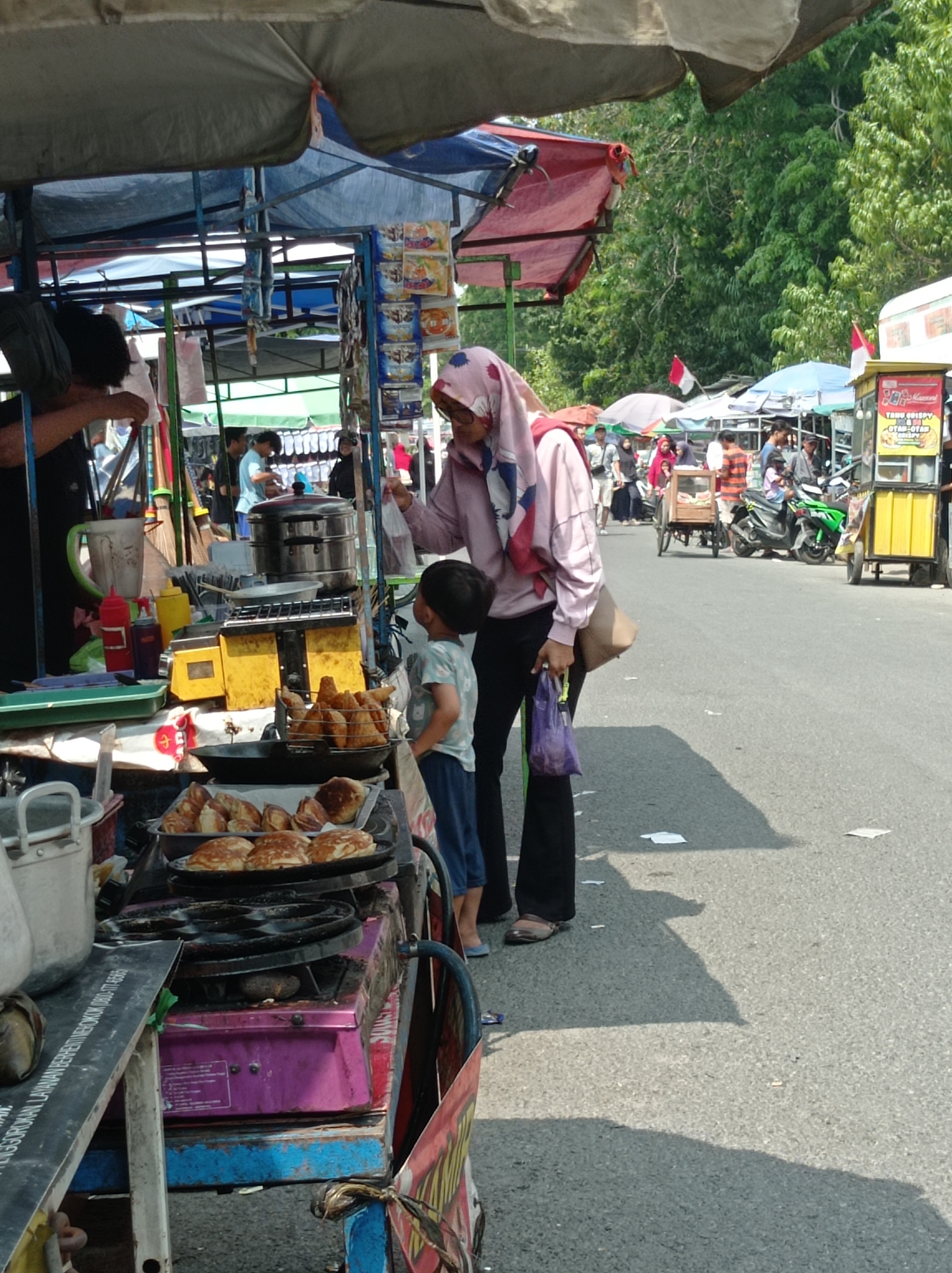 This image depicts a street scene in Tegal, Indonesia, likely a market or roadside stall area. In the foreground, a food cart is prominent. A woman wearing a pink hijab and a light pink jacket is interacting with the cart, holding a purple plastic bag. A young boy in a light blue t-shirt and dark pants stands beside her, looking at the food. The cart displays various food items, including fried food in a basket and pancakes on a plate. There are cooking utensils and containers on the cart, including a yellow scale and a multi-tier steamer. The background shows other market stalls, people walking, and a bright, sunny day with trees in the distance. A red and white flag is visible, possibly indicating Indonesia's national flag. The environment appears to be a paved road.