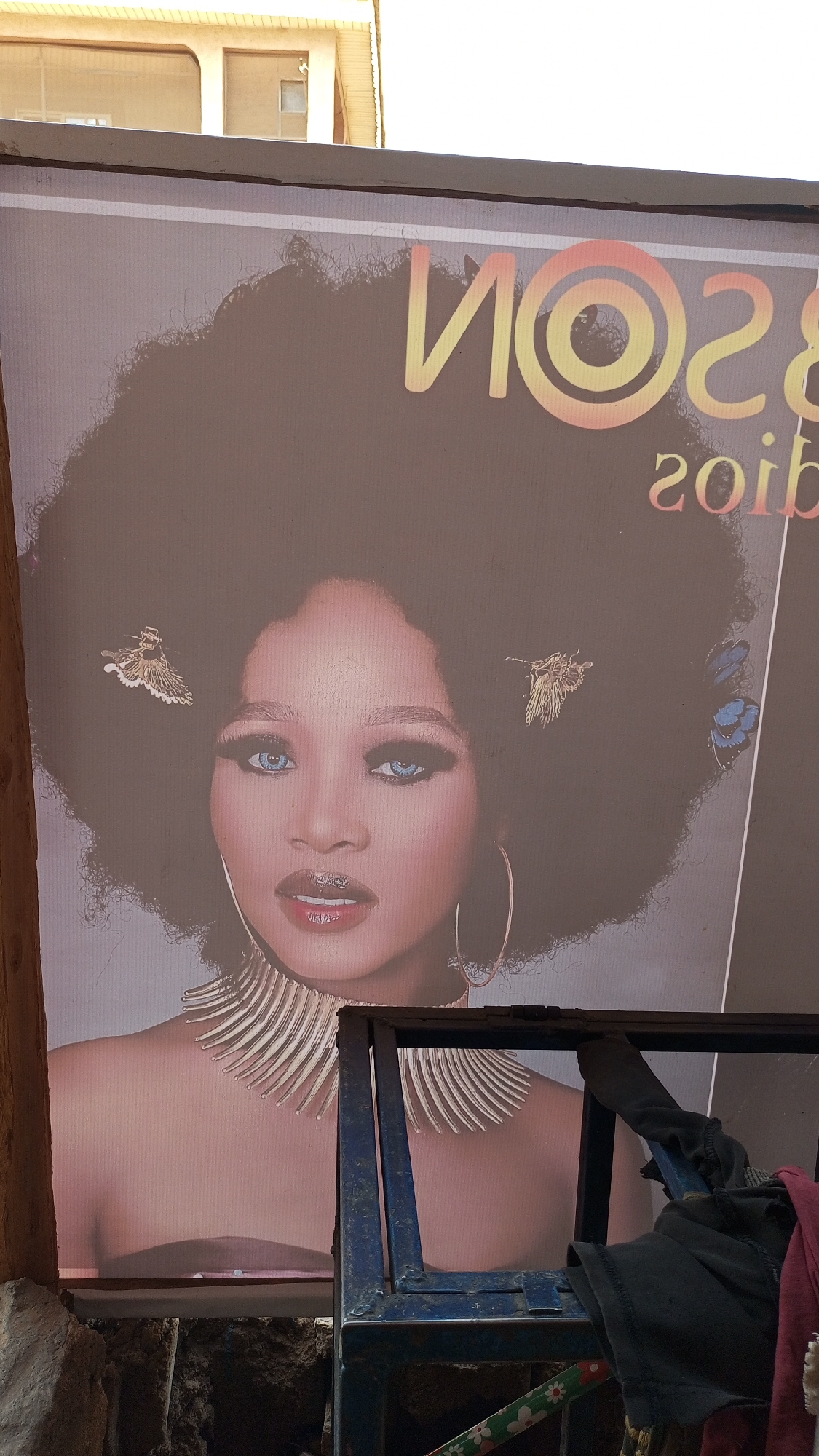 A close-up shot shows a poster or sign for "NOS28", which appears to be an advertisement for a fashion or beauty-related event or brand in Yelwa, Nigeria. A woman with dark skin, an afro hairstyle, and strikingly light-colored eyes is featured prominently. Her hair is adorned with accessories resembling insects or butterflies. She wears a large, elaborate necklace resembling a spine or ribcage and large hoop earrings. The poster is mounted on a surface, and a blue metal frame or structure partially obscures the lower portion of the poster. An unidentifiable item, potentially clothing, hangs on the frame. The surrounding environment is blurry, indicating the poster is the main subject. The picture is taken in daylight, suggesting an outdoor setting.