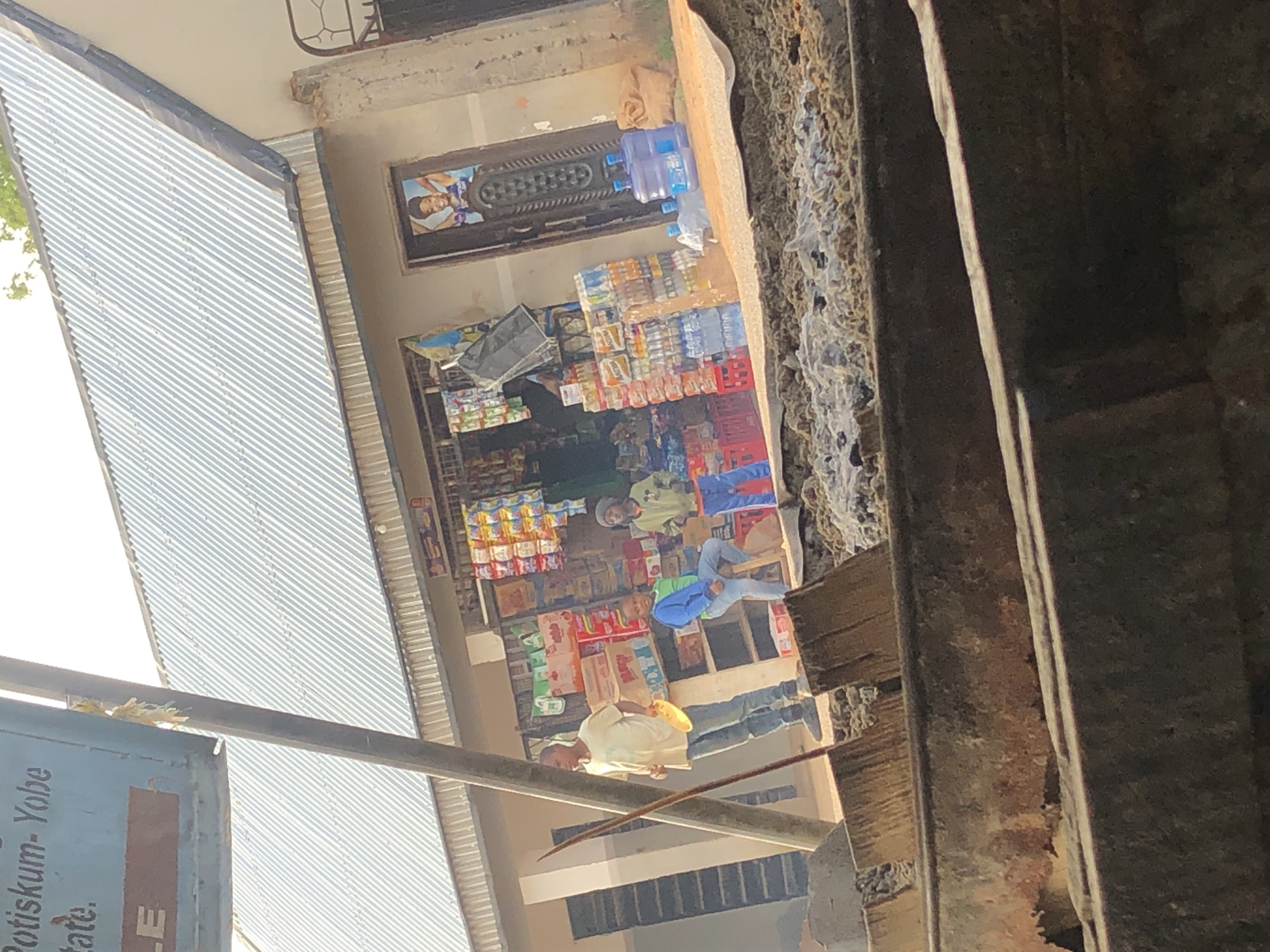 The image depicts an outdoor scene in Jos, Nigeria, likely in front of a small shop or market stall. In the foreground, a large, rough, concrete structure with a dirty, uneven surface is visible, suggesting a construction site or a damaged area. Beyond this, a group of people are gathered. One man, wearing a light-colored t-shirt and jeans, stands with a yellow circular object in his hand. To his right, a younger person in a blue jacket and green shirt is seated on a wooden stool. Further to the right, another young person in a green jacket and orange pants stands. Behind them, the shop is stocked with various goods, including stacks of bottled water and what appear to be packaged snacks and drinks. A corrugated metal roof covers the structure. A framed picture of a smiling young woman is displayed on the door of the shop. The overall impression is of a busy, everyday scene in a Nigerian city.