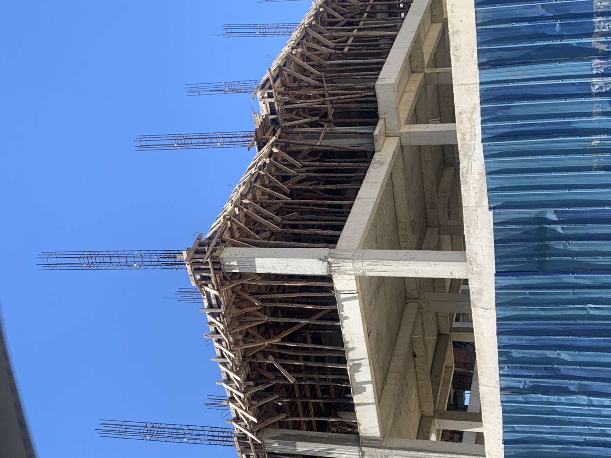The image shows a building under construction against a clear blue sky. The building's structure is primarily composed of reinforced concrete pillars and beams. Wooden scaffolding and supports are visible, particularly on the upper levels. Metal rebar extends from the top of the structure. Blue sheeting or cladding partially covers a section of the building. The location is Dar es-Salaam, Tanzania.