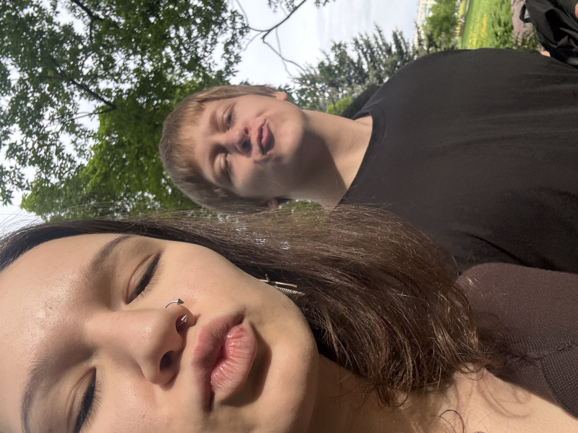 The image captures a playful outdoor selfie of two young individuals, presumably taken in Saint Petersburg, Russia. The photo is rotated counter-clockwise, showing the subjects looking towards the top-left of the frame.
In the foreground, a young female has her face angled towards the camera, with eyes closed and lips slightly pursed in a pout. She has dark brown hair, fair skin, and a prominent silver horseshoe barbell piercing with spikes in her septum. A long, thin metallic earring is visible on her left ear. She appears to be wearing a dark brown top.
Behind her, a young male's face is slightly out of focus but clearly visible. He also has his lips pursed in a playful pout, with eyes partially closed. He has light brown or blond hair and is wearing a black t-shirt. A black item, possibly a backpack, is visible on the far right edge behind him.
The background features a vibrant green canopy of deciduous trees against a bright, possibly overcast or hazy, sky, indicating an outdoor setting like a park or garden. Hints of distant green grass or a field and light-colored buildings can be seen further in the background, suggesting an urban park setting consistent with locations found in Saint Petersburg. The bright natural light suggests it is daytime. The overall mood of the image is casual, relaxed, and affectionate. There is no visible text in the image.