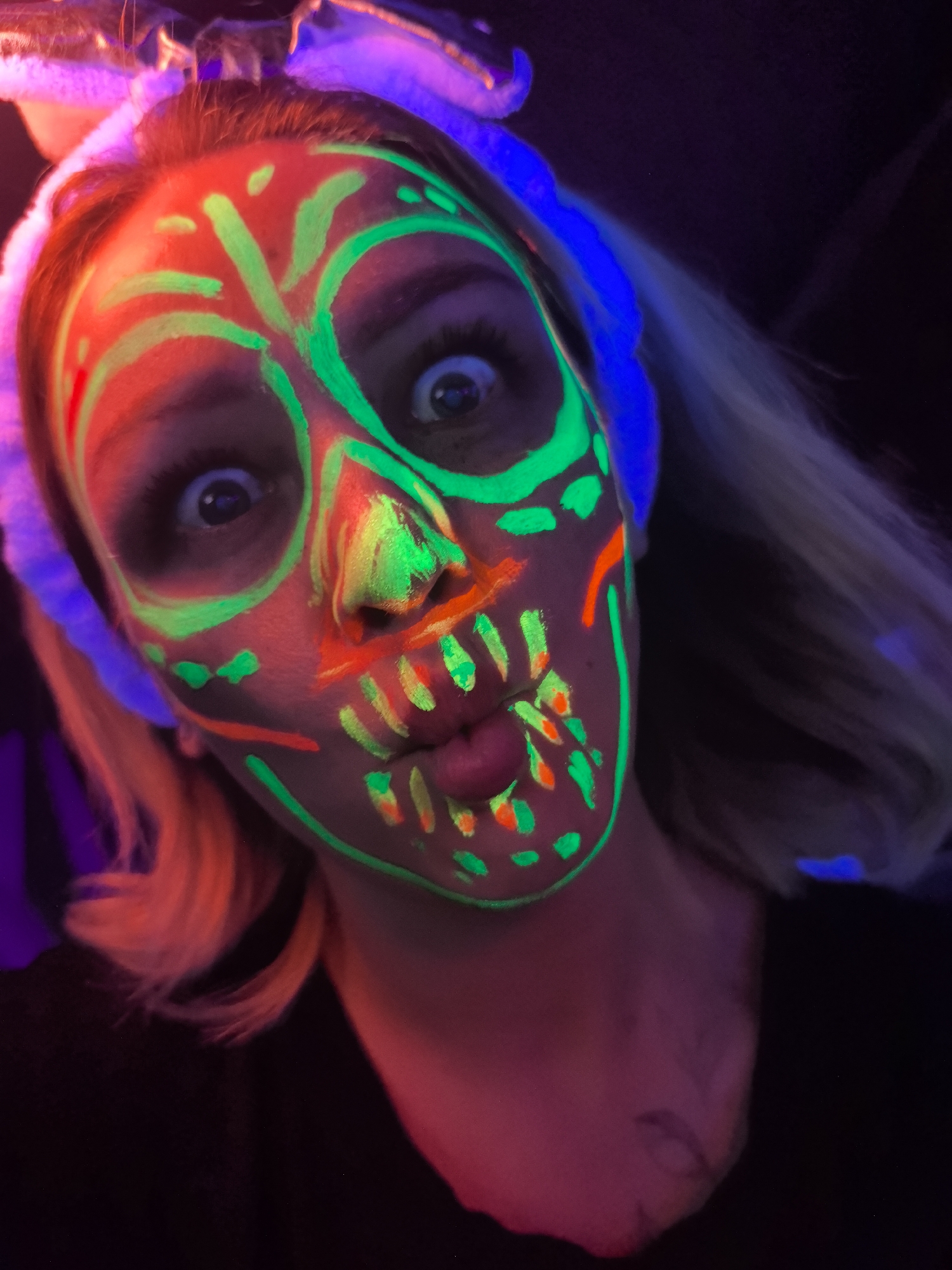 A woman with fluorescent face paint resembling a skull is looking directly at the camera. The face paint is primarily green and orange. She has wide eyes and pursed lips. She is wearing a white, soft headband and has light-colored hair. The background is dark, with hints of purple and blue lighting. No location information can be determined from the image.