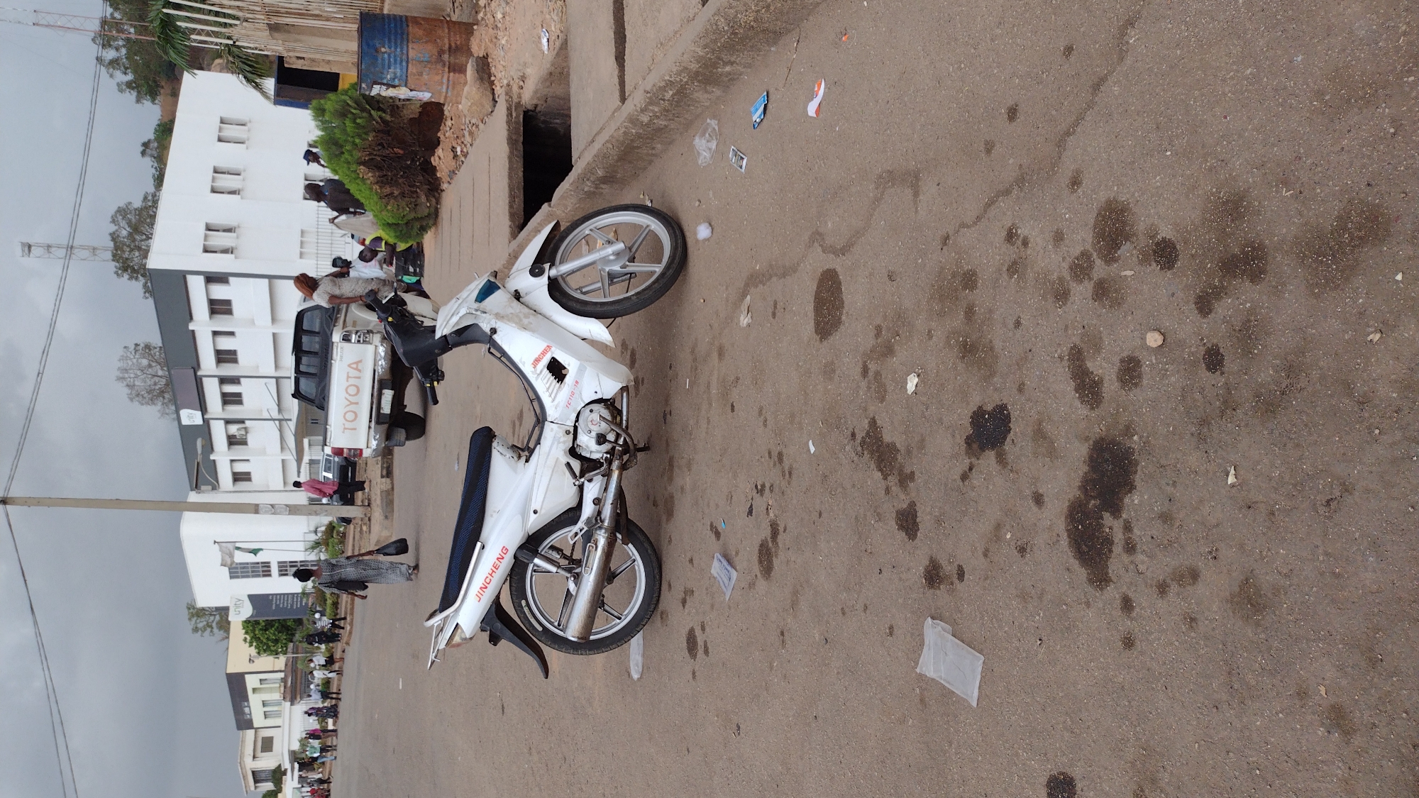 The image captures a scene in Jos, Nigeria, showing a white Jincheng motorcycle lying on its side on a paved road. The motorcycle appears damaged at the front. The road surface has dark fluid stains and scattered debris. In the background, there's a white Toyota pickup truck, and groups of people are gathered near buildings and a utility pole. The sky is overcast.