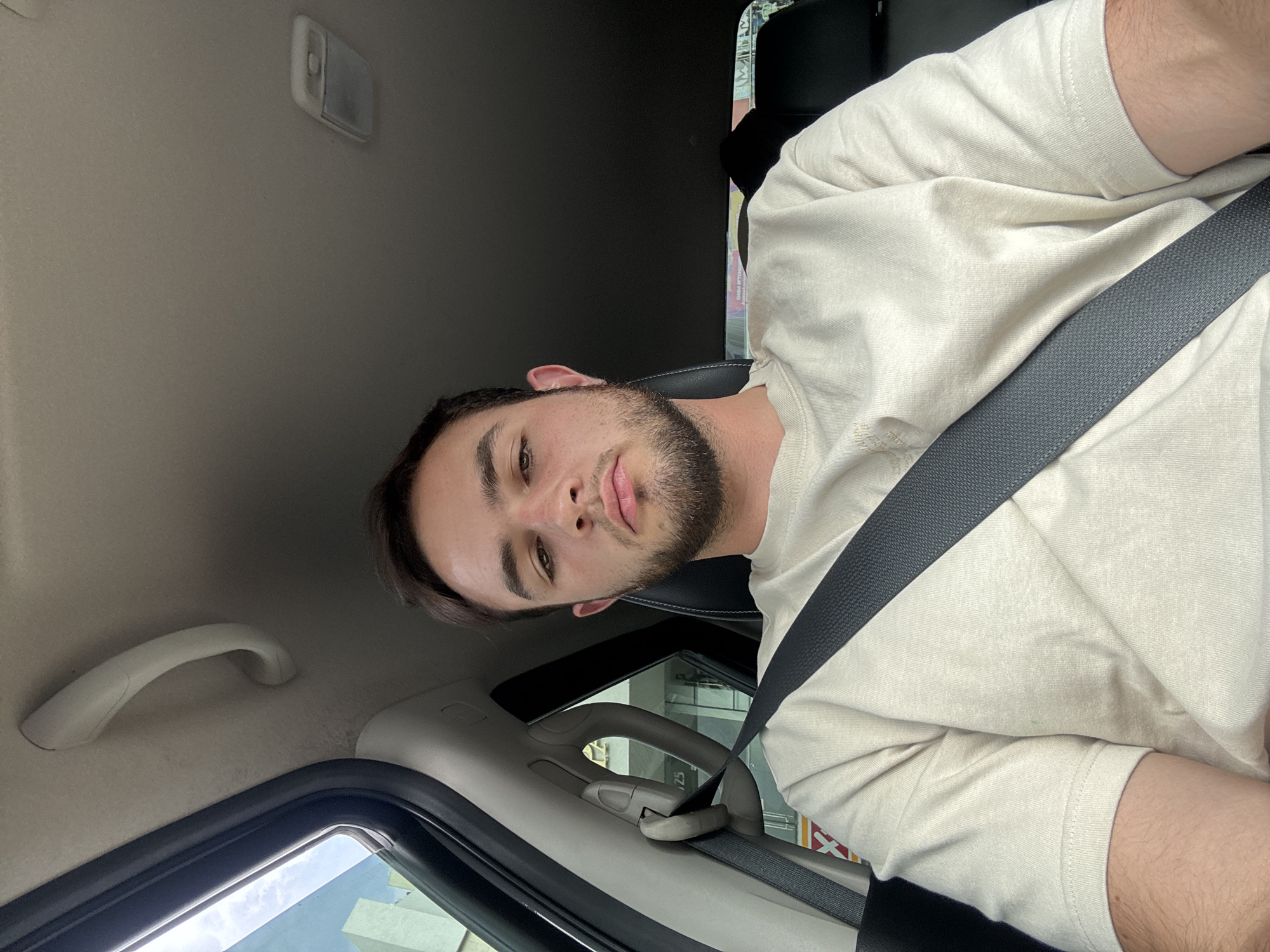 A medium, canted, indoor shot depicts a young man wearing a seatbelt in what appears to be a car. He is facing the camera, which is positioned at a slightly higher angle. The interior of the vehicle is visible, including the roof, window frames, grab handle, and part of the driver's seat. Through the window, there is a view of the outside, including a building. There is no specific information in the image to confirm or deny its location as San Bernardino Tlaxcalancingo, Mexico.