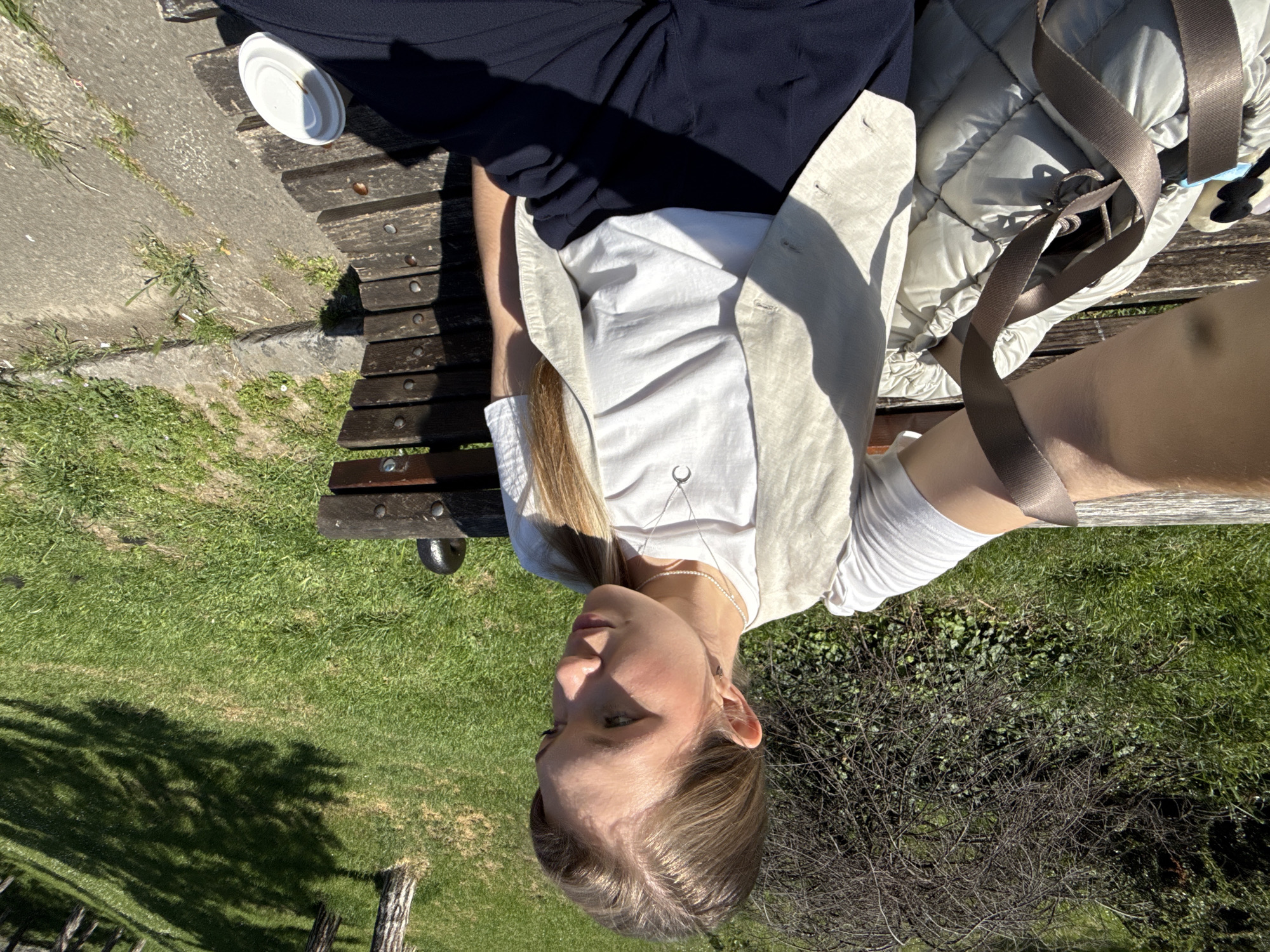 A young, light-skinned woman sits on a wooden bench in a park on a sunny day. She wears dark trousers, a white shirt, and a beige waistcoat. A light-colored padded jacket and a white cup rest on the bench beside her. The park contains grass and a leafless bush and is in Belgrade, Serbia.