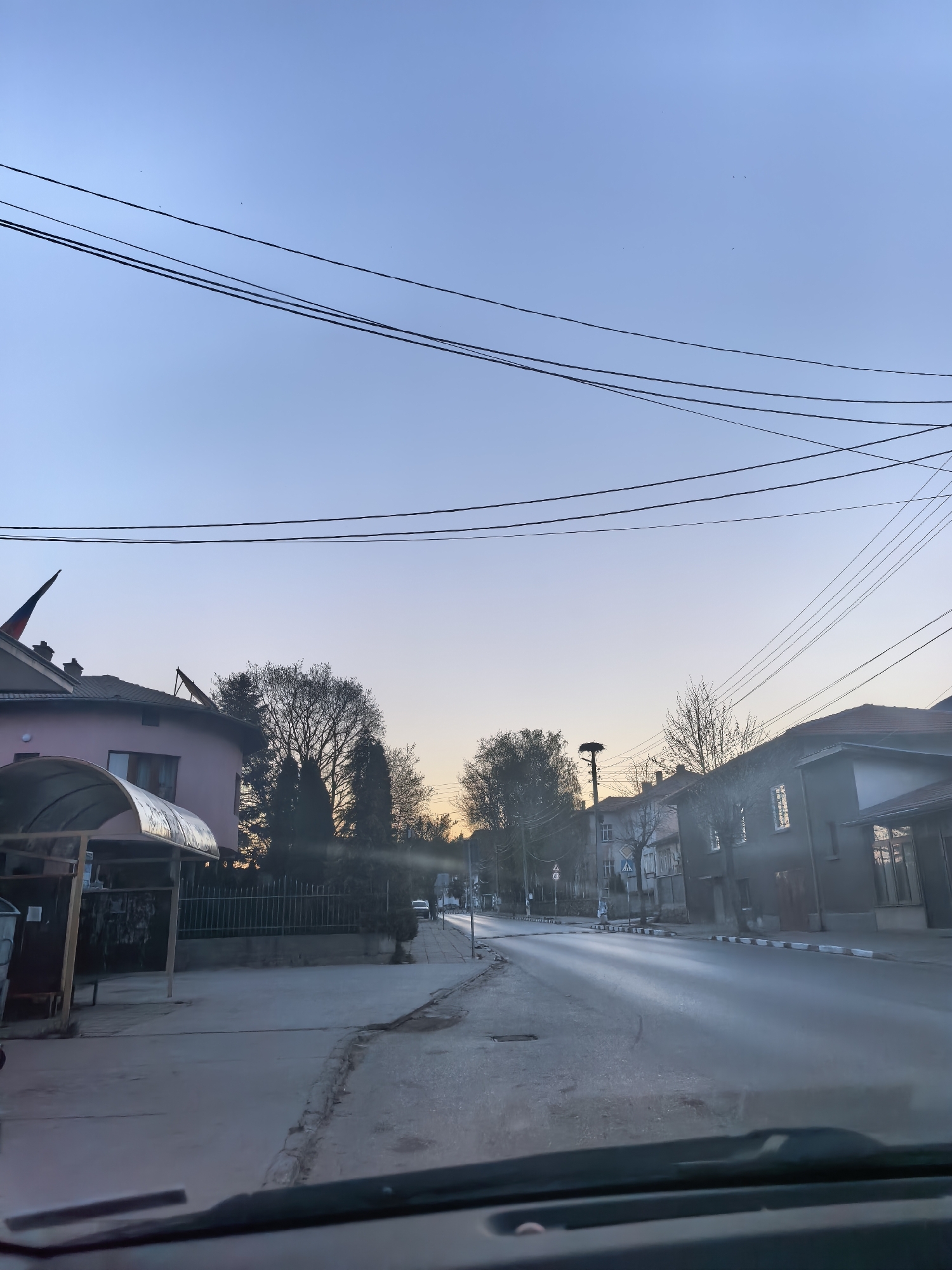The image shows a street in an urban environment, likely a small town, during dawn or dusk. The road is paved and leads away from the camera, with buildings on either side. The building on the left has a pink facade and an arched metal structure next to it. The building on the right is a darker gray color. Overhead are several electrical or telephone wires stretching across the sky. In the distance, between the buildings, are trees and what appears to be a tall pole structure with a platform or nest-like feature at the top. The sky is a gradient of colors, transitioning from a light blue at the top to an orange-yellow near the horizon. The image appears to be taken from inside a vehicle, with the dashboard visible at the bottom of the frame. There are street signs visible on the right side of the road in the distance. No people are visible in the image.