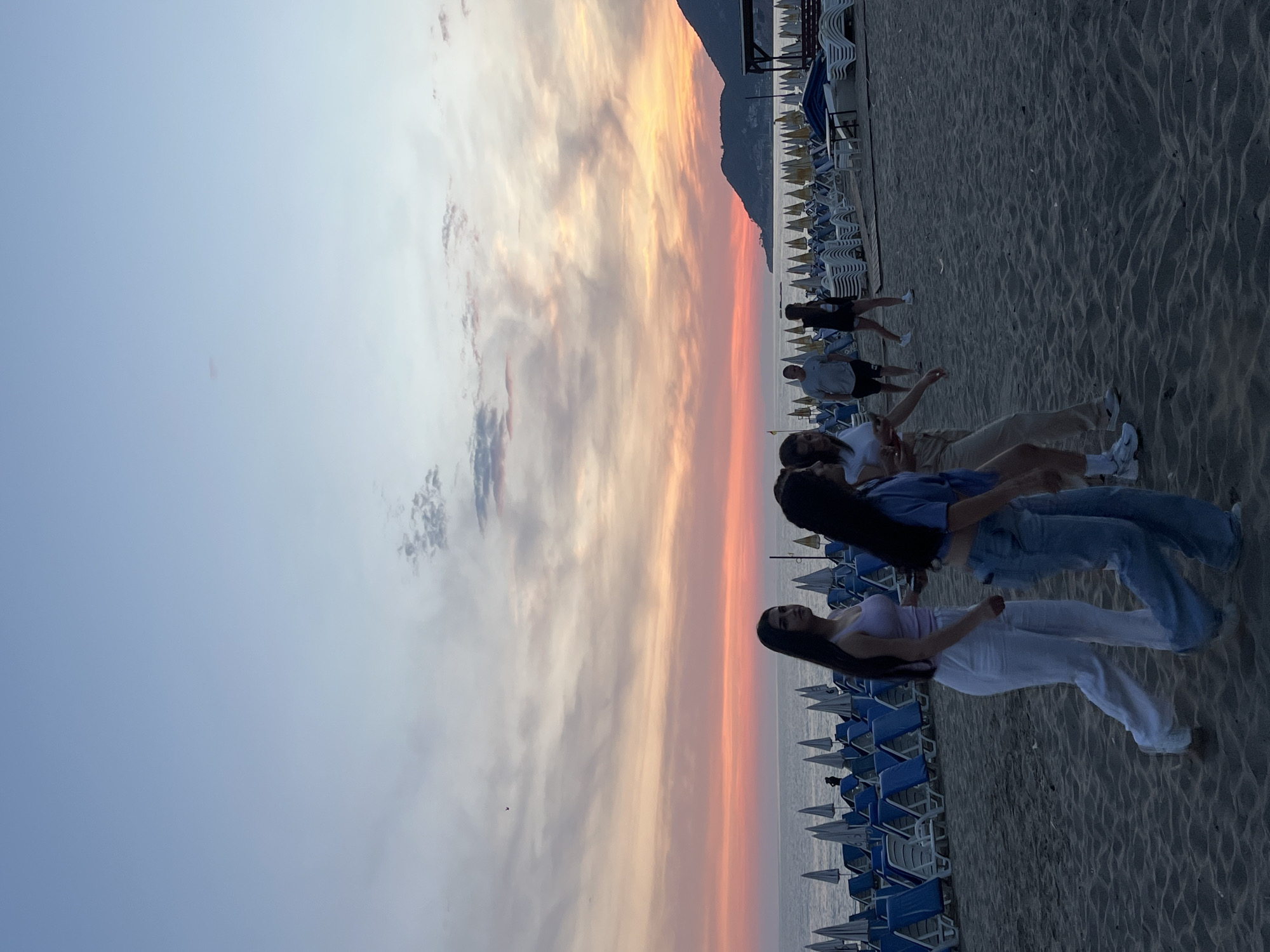 The image depicts a sunset scene on a beach in Alanya, Turkey. The sky is a blend of orange, pink, and light gray hues, indicating sunset. A mountain range is visible in the background.
A group of people is standing on the sand, with some lined up closer to the camera. From front to back, the people include: a woman with long dark hair, wearing a light purple top and white pants; a person wearing a blue shirt and blue jeans; and other people in lighter colored clothing.
Numerous blue and white beach chairs and umbrellas are lined up on the sand, extending into the distance. The beach is sandy, and the sea is visible in the background. Some other people are walking further down the beach in the distance.