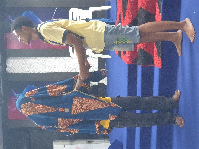 The image portrays an indoor scene in Onitsha, Nigeria, featuring two individuals standing barefoot on a blue carpeted floor, engaging in a two-handed handshake. The person on the left is largely obscured by a blue garment with a geometric pattern of orange, red, and white, resembling traditional African fabric, worn over a yellow shirt and dark trousers. The person on the right is a young male, dressed in a yellow polo shirt with a blue collar and a visible lanyard, and light blue denim shorts featuring a green and white checkered patch on the left thigh. This individual faces left, looking upwards and slightly left with a subtle facial expression. Behind the subjects, a blue carpeted stage with two visible steps is present, topped by a shaggy red and black patterned rug. The background wall consists of distinct panels, including a white panel with a repeating star-like geometric pattern and darker panels with subtle designs. Two white plastic chairs are visible in the background to the right. A white cabinet or appliance is partially visible on the far left.
