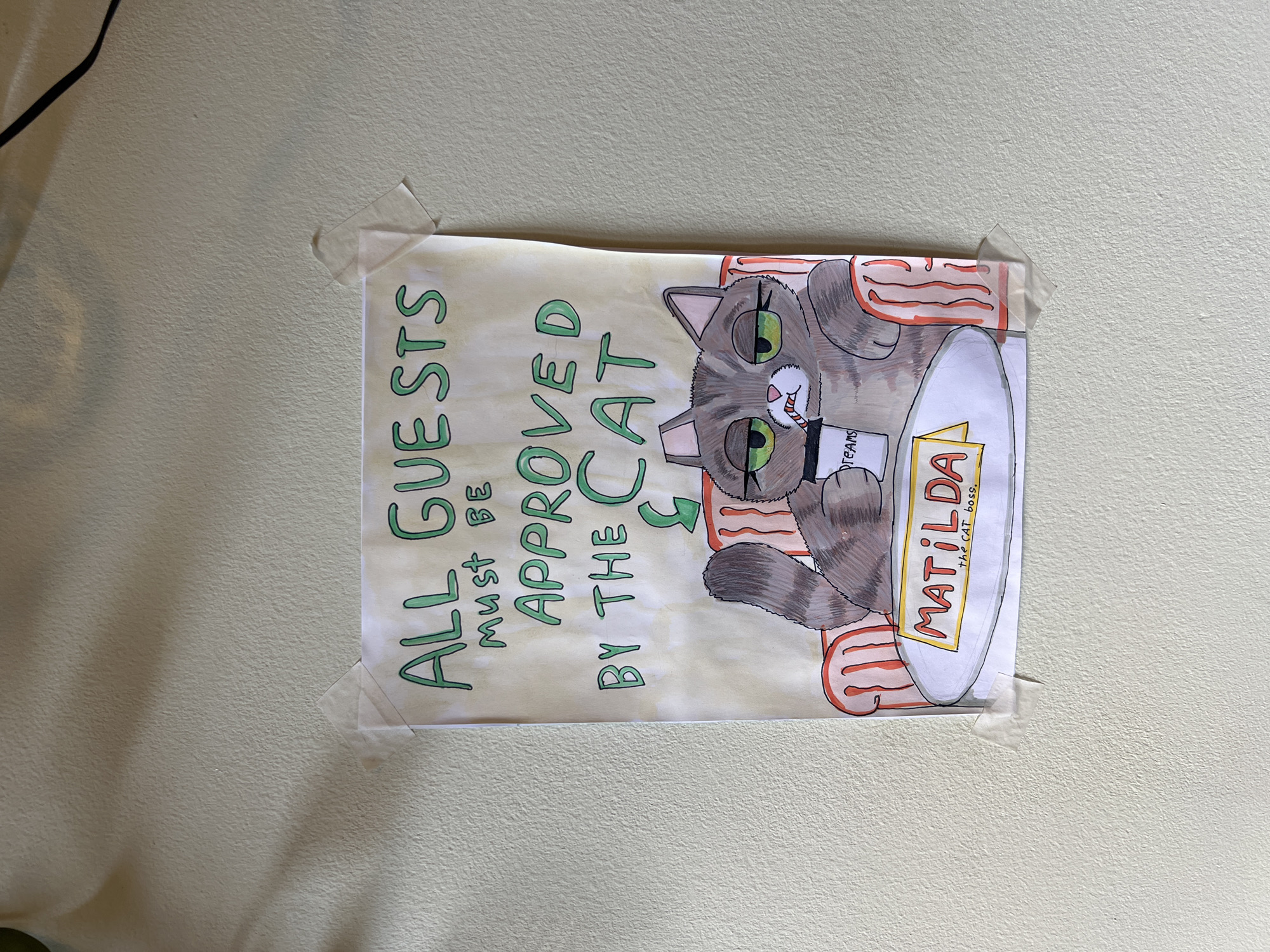 A drawing is attached to a pale beige wall with transparent tape at each corner. The drawing features a cartoon cat sitting on a red-and-white striped bed, holding a magazine titled "DREAMS". The cat has green eyes, grey and white fur, and a stern expression. In bold, green letters to the left of the cat, the words "ALL GUESTS MUST BE APPROVED BY THE CAT" are written. The cat bed is labeled "MATILDA the CAT boss."
The location in the city of Budva, Montenegro, cannot be determined based on the provided image.