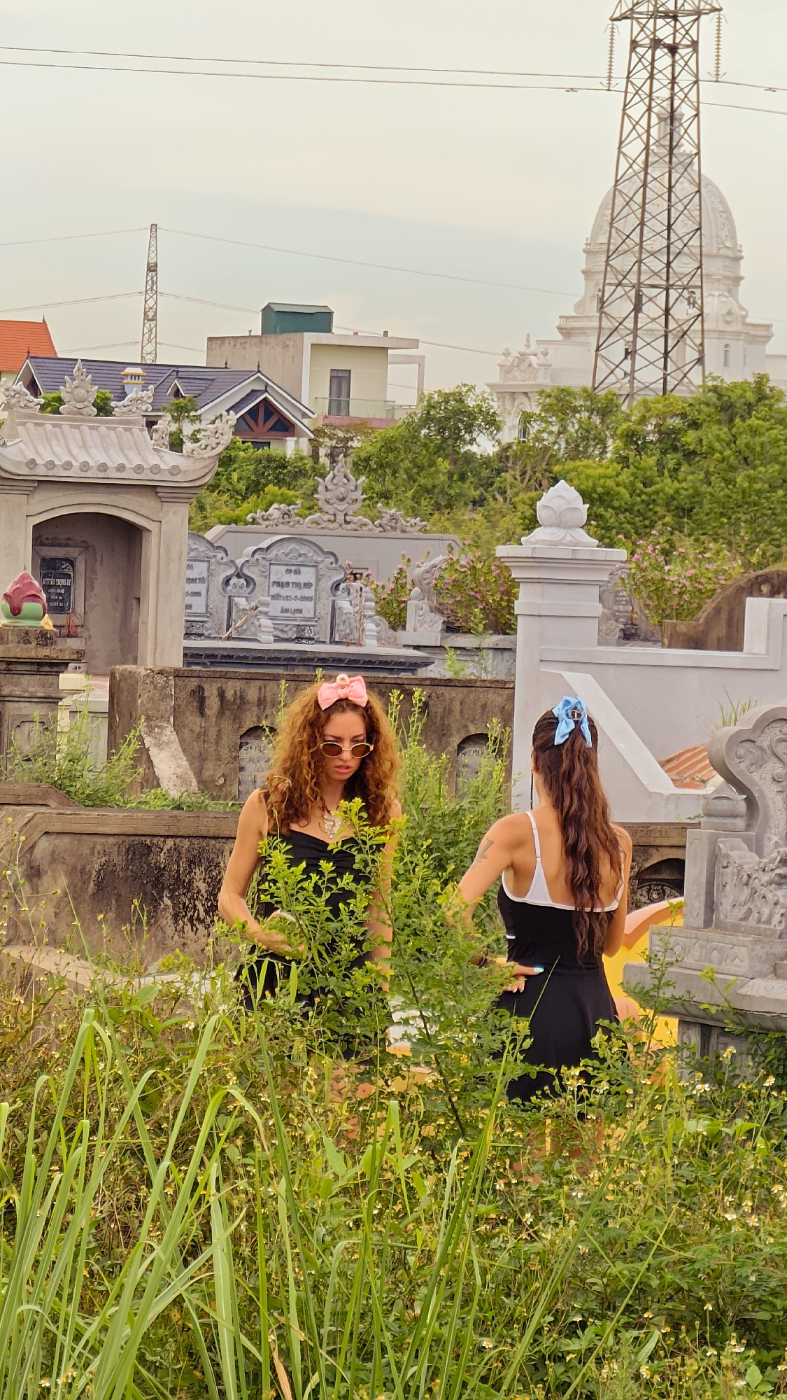 The image depicts two women standing amidst overgrown vegetation within a cemetery in Hoa Lư Ward, Vietnam. The woman on the left, with curly brown hair adorned with a pink bow, wears sunglasses and a black dress. The woman on the right, facing away from the camera, has long dark hair tied with a blue bow and wears a black and white dress. Behind them, traditional Vietnamese cemetery structures and elaborate tombs are visible, interspersed with lush greenery. In the background, a large industrial transmission tower and a dome-shaped building are prominent against an overcast sky. The overall scene is one of a cemetery undergoing natural overgrowth.