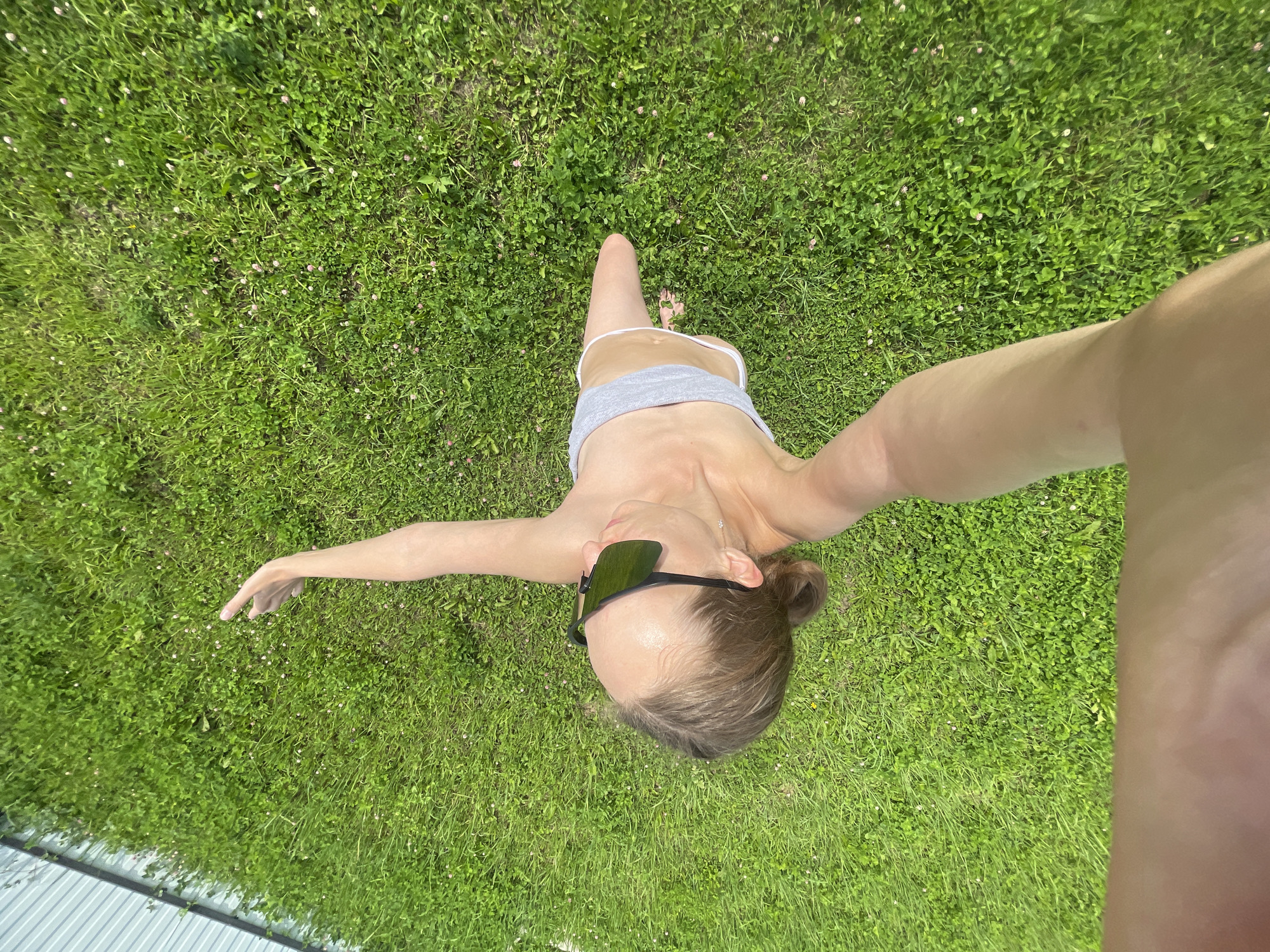 This is a top-down, wide-angle shot of a woman lying on a grassy lawn in Подшивалово, Russia. The woman is wearing sunglasses, a grey tank top, and light-colored shorts. Her arms are outstretched to the sides. The grass is green and dotted with small pink flowers. In the foreground, the edge of a white corrugated metal fence is visible. The lighting suggests it is a sunny day, likely in the afternoon, given the strong shadows. The woman's pose and the bright outdoor setting evoke a sense of relaxation and enjoyment of the outdoors.