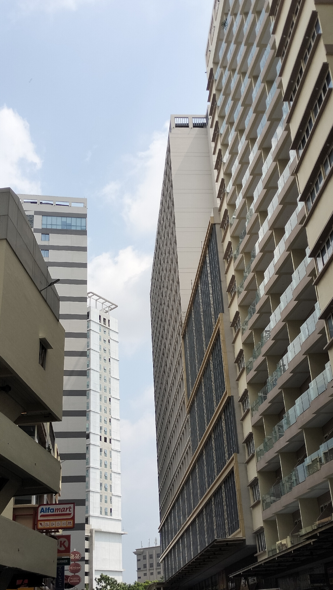 The image captures a view looking up at several high-rise buildings under a bright, cloudy sky in Batam, Indonesia. The foreground features the side of a beige and gray building with a narrow, dark horizontal window. Below this, a red and white sign for "Alfamart" is visible, along with other smaller signs.
To the left and slightly further back, a tall, slender building with alternating horizontal bands of gray and white is prominent. This building has numerous small windows.
In the center, another tall, white building rises, also featuring many windows and a modern, angular rooftop structure.
To the right, and dominating the right side of the frame, is a much wider building with a repeating pattern of balconies, each enclosed by a glass railing. The facade of this building is a light beige color, with dark brown trim around the windows and balconies.
Further in the distance, a smaller, darker building with a grid-like facade can be seen. The overall scene depicts an urban environment with a concentration of modern architecture.