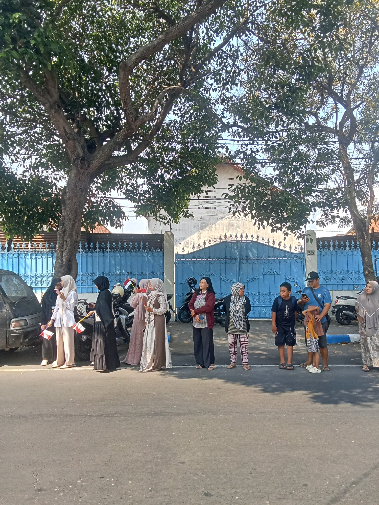 A group of people are standing on a paved street in front of a blue gate. The gate is ornate and appears to be made of metal. Behind the gate, a building with white walls and a red roof is visible. Large trees with green foliage are overhead, casting shadows on the ground. The people are dressed in a mix of casual and traditional clothing. Several individuals are wearing hijabs. Some are holding small Indonesian flags. A black car and several motorcycles are parked nearby. The scene appears to be in an urban or suburban area, likely a street in Jombang, Indonesia. The time of day is daytime, as indicated by the bright sunlight and shadows.