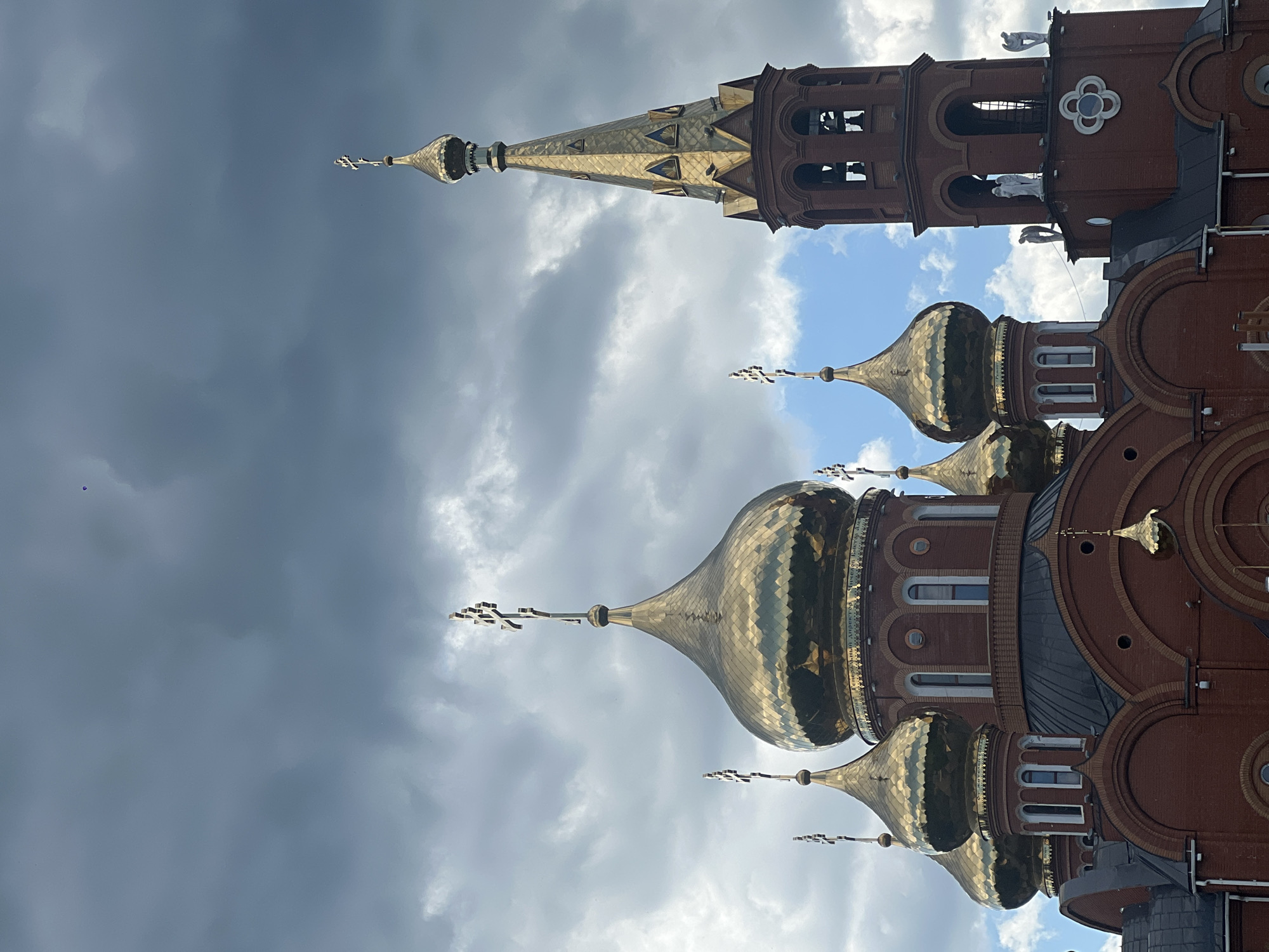 This image captures a portion of the Orthodox Church of St. Nicholas in Chaikovsky, Russia, viewed from a slightly low angle against a dramatic sky. The church features a prominent red brick construction. In the foreground, multiple onion-shaped golden domes are visible, each topped with an ornate Orthodox cross. The largest dome, centrally located among the group, displays a patterned, reflective golden surface and has an inscription in Cyrillic script around its base, partially visible as "ГЕНЕРАЛЬНЫЙ ДИРЕКТОР".
To the right, a tall, slender bell tower rises, constructed from the same red brick and capped with a golden, spire-like dome, also adorned with a cross. The bell tower has arched openings revealing bells within. On the flat roof sections of the bell tower's lower structure, three white figures resembling angels are positioned. The sky behind the church is largely overcast with heavy, dark grey clouds, though patches of brighter blue sky are visible, particularly towards the lower right. A small, dark, roughly circular object, possibly a balloon or drone, is visible in the upper right portion of the sky, near the edge of the clouds. Another minute, dark, irregular object is present in the upper left sky.