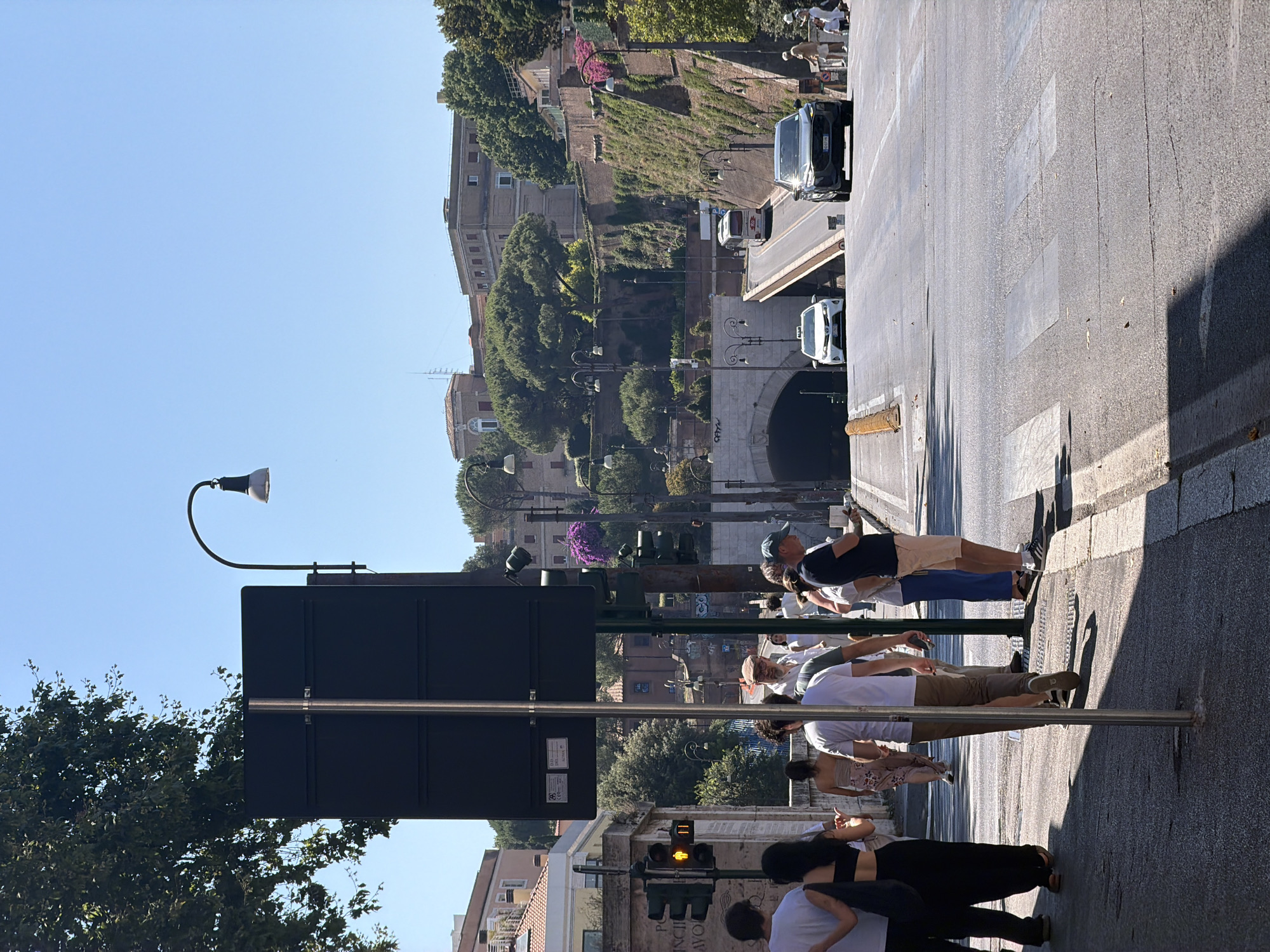 In an urban environment with clear blue skies, a street scene unfolds. In the foreground, a pedestrian crossing is marked on asphalt, with shadows from nearby objects cast across it. Several individuals are visible on the sidewalk and at the crossing. A traffic light displays a yellow pedestrian symbol. To the left, a large, dark rectangular sign stands vertically.
The middle ground features a road leading towards a large arched tunnel entrance. Cars, including a white taxi and a dark SUV, are traveling on the road. A white bus is seen on the incline leading to the tunnel. The surrounding area is characterized by a retaining wall covered in vegetation and stone, with buildings and trees visible in the background. The overall impression is a sunny day in a city with historical architecture and modern infrastructure.