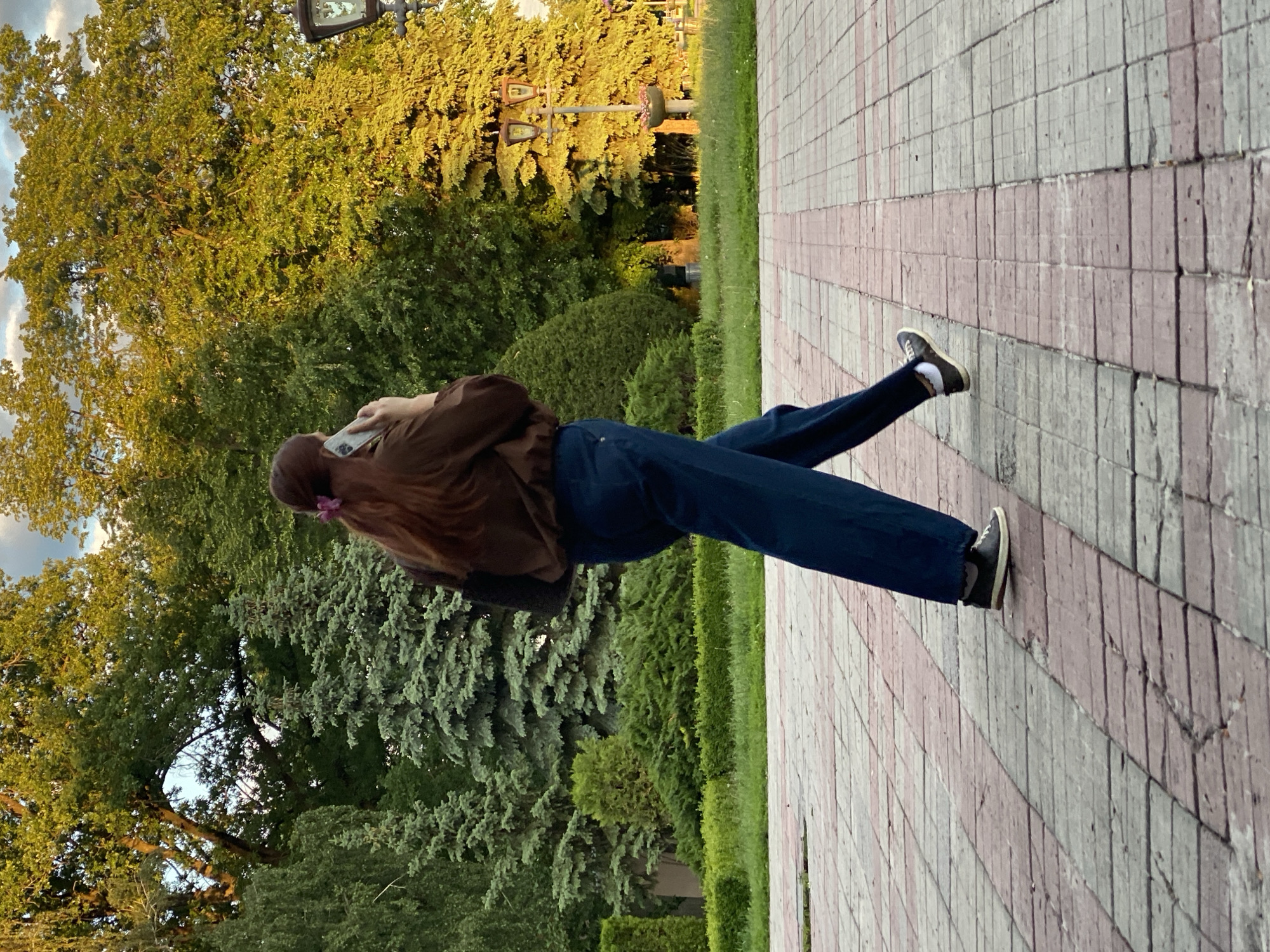 The image depicts an outdoor scene, likely a park or public pathway, in Poltava, Ukraine, bathed in the warm light of late afternoon or early evening. A person, viewed from behind, stands on a paved path, engaged with a smartphone held up to their ear or facing them.
The subject is wearing a brown long-sleeved top or jacket, dark blue jeans, and dark sneakers with white socks. A pink hair tie or accessory is visible in their long, dark hair. They appear to be casually standing or slowly walking along the path.
The setting is characterized by abundant greenery, including tall deciduous trees with bright green leaves, and darker coniferous trees. The light suggests the "golden hour," casting a warm glow on the foliage. The paved path features a distinct pattern of alternating light grey and reddish or pinkish rectangular blocks, showing some signs of wear. Beyond the path, there is green grass and well-maintained shrubbery. In the background, a streetlamp with two fixtures is visible, and faint shapes among the trees might indicate playground equipment or distant structures. The sky, partially visible, shows a few light clouds. No legible text is visible in the image.