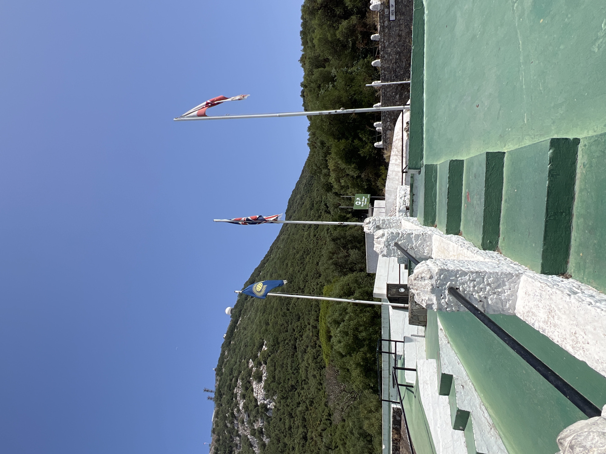 The image shows a view of the Great Siege Tunnels in Gibraltar. In the foreground, there are green-painted steps leading up to a viewing platform. The steps are bordered by white stone walls with black railings. In the background, there are three flags flying from poles. The left flag is blue with a yellow emblem. The middle flag is the Union Jack. The right flag is red and white and appears to be a British flag. Behind the flags, the green, tree-covered slope of the Rock of Gibraltar is visible, with some communication towers and a white dome-shaped structure on its summit. The sky is clear and blue. A sign with a green arrow and the words "Great Siege Tunnels" is visible between the blue flag and the Union Jack.