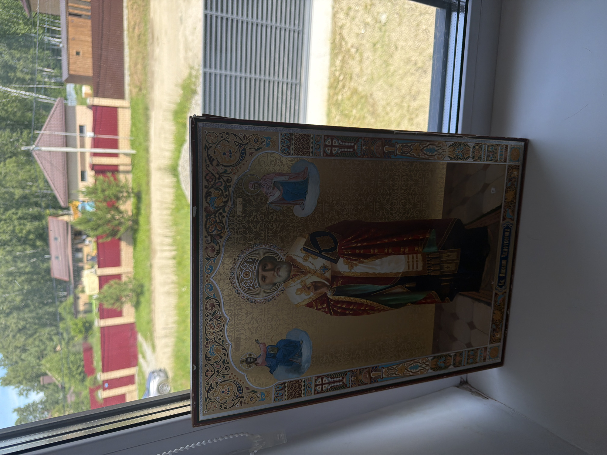 The image shows an icon of Saint Nicholas, a religious figure, placed on a windowsill. The icon is framed and features intricate gold detailing. Behind the icon, through a window, a suburban landscape is visible, with houses, fences, trees, and a car. The lighting suggests it is daytime. The icon appears to be a replica of a religious painting. It is not possible to determine the exact location within Malinovka, Russia, from the image alone.