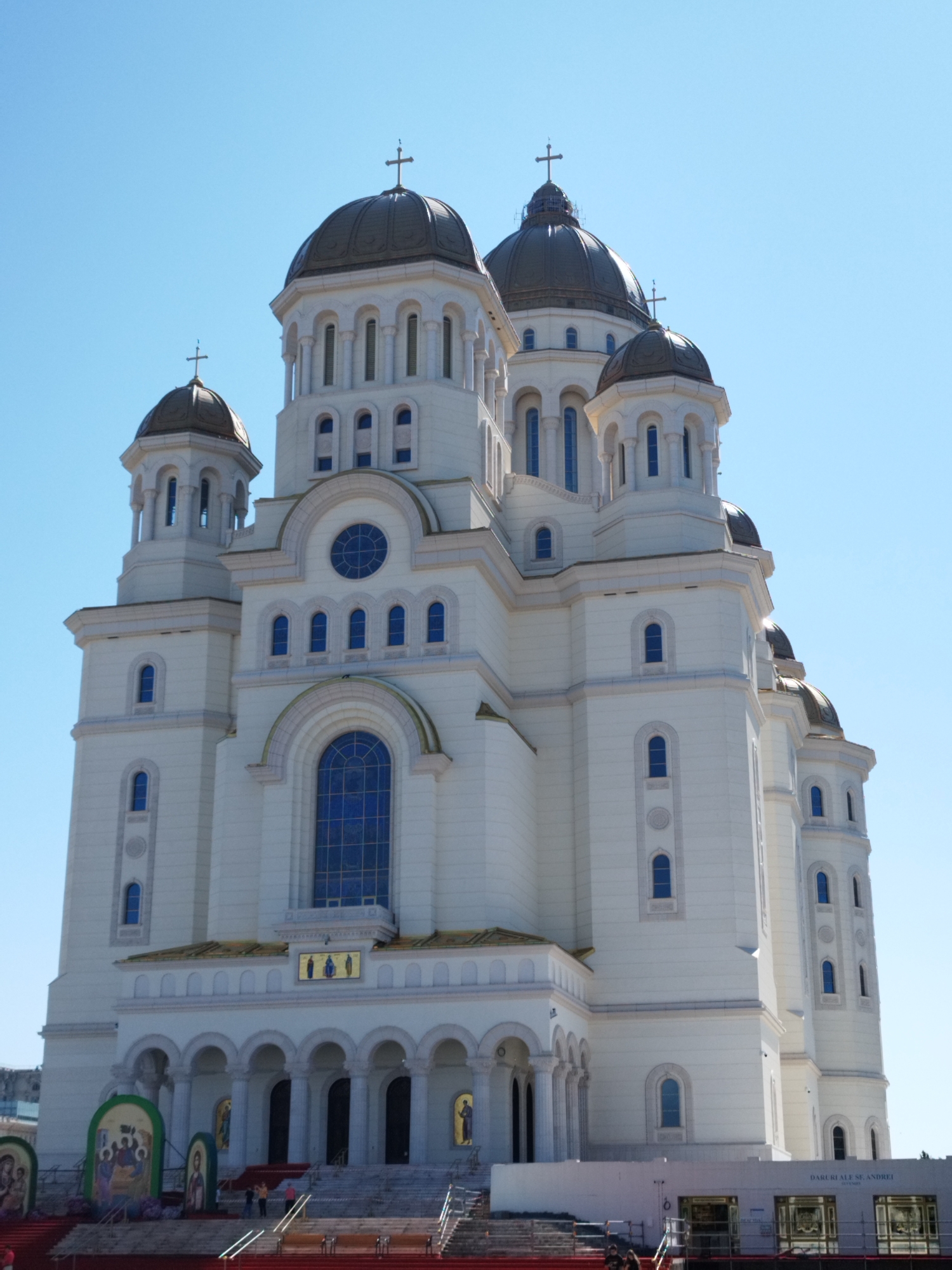 The image shows the exterior of the Metropolitan Cathedral of Saint Constantine and Helena in Bucharest, Romania, under a clear blue sky. The large, white cathedral features multiple domes, the largest of which is dark and topped with a cross. The architecture is ornate, with numerous arched windows, columns, and decorative elements. Smaller domes and towers are visible on the sides and at different levels of the building. The facade is primarily white with gold accents and blue windows. In the foreground, there are red stairs leading up to the cathedral's entrance, which is framed by arches and columns. Several religious icons are displayed on stands in front of the stairs, and a few people are visible on the stairs and in front of the cathedral's base, some appearing to be tourists. The overall scene conveys a sense of grandeur and religious significance.