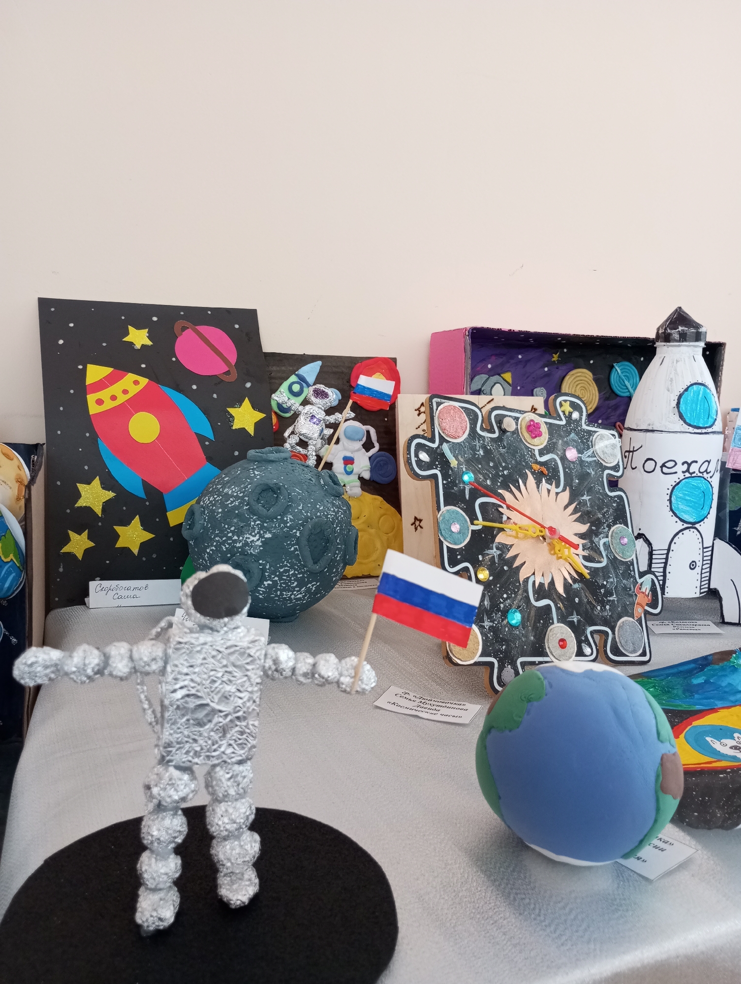 The image shows a display of space-themed crafts, likely created by children, on a light-colored surface. The crafts include: a rocket and planets on a black background with yellow stars; a textured sphere representing a moon or planet; tin foil astronauts holding flags; a Russian flag; a puzzle-piece clock with planets and space-related details; a rocket; a diorama; and a model of Earth. A white wall is visible in the background. The location is unknown.