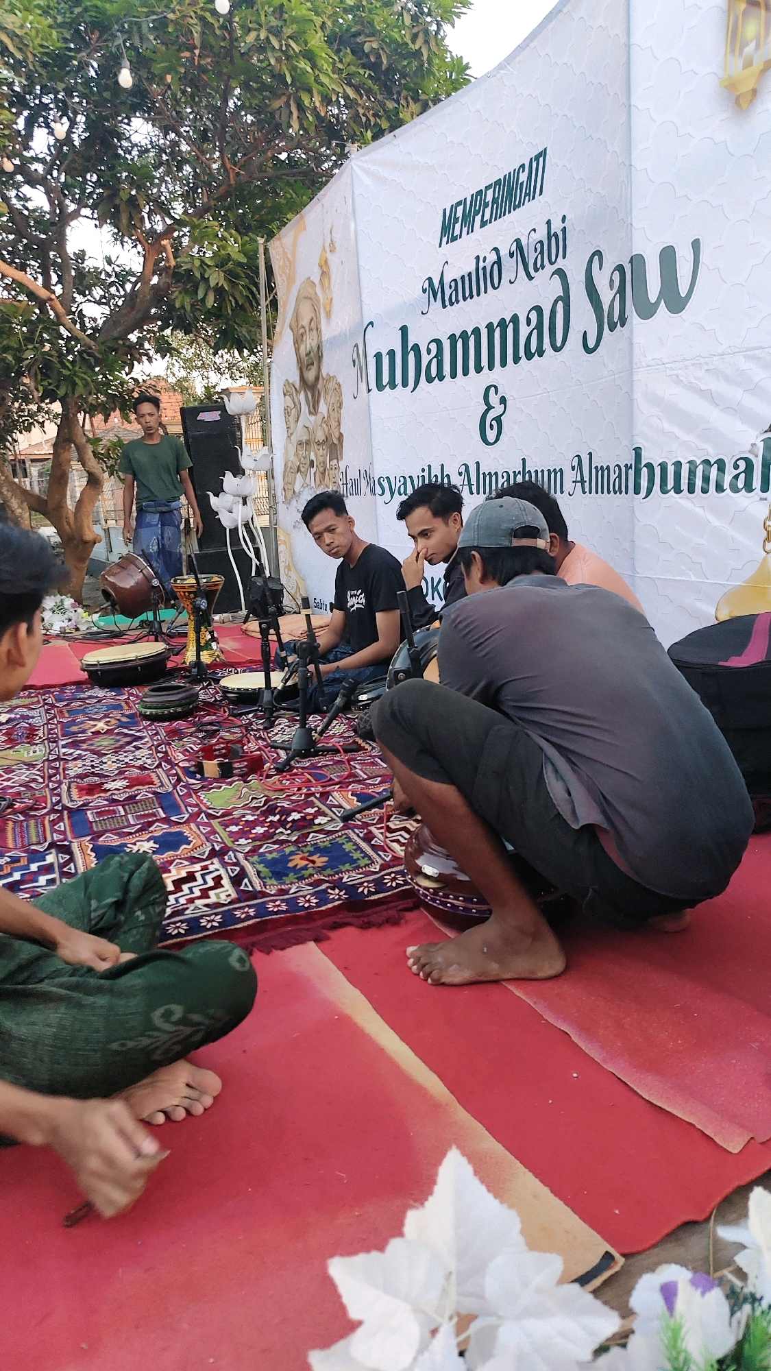 In Galis, Indonesia, a group of young men are gathered outdoors for an event. A large white banner with green text serves as a backdrop. The banner reads "MEMPERINGATI Maulid Nabi Muhammad Saw & Haul Masyayikh Almarhum Almarhumah." Several young men, dressed casually, are seated or crouching on a patterned rug and a red carpet. Some are looking towards the camera, while others are engaged in conversation or preparing for a performance. Musical instruments, including drums and possibly other percussion, are arranged on stands. A speaker system is also visible. The environment suggests a community gathering or religious celebration taking place during daylight hours, with trees and foliage in the background.