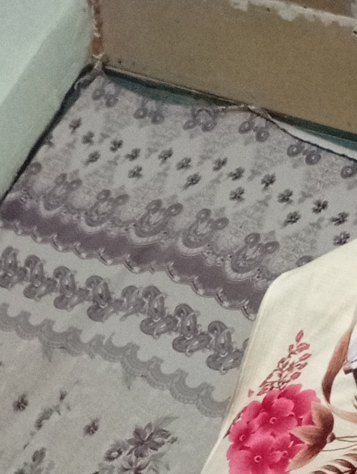 The image shows a close-up view of a patterned fabric, likely a bedspread or blanket, with a floral design. The fabric is predominantly light gray with darker gray and purple patterns. The pattern includes intricate paisley-like shapes and smaller floral motifs. In the lower right corner, a portion of another fabric is visible, featuring a large, vibrant pink flower and some brown branches. The background appears to be a wall and a corner, suggesting an indoor setting. There are no people visible in the image, and no specific location in Isfayram, Kyrgyzstan can be identified from the visual information.