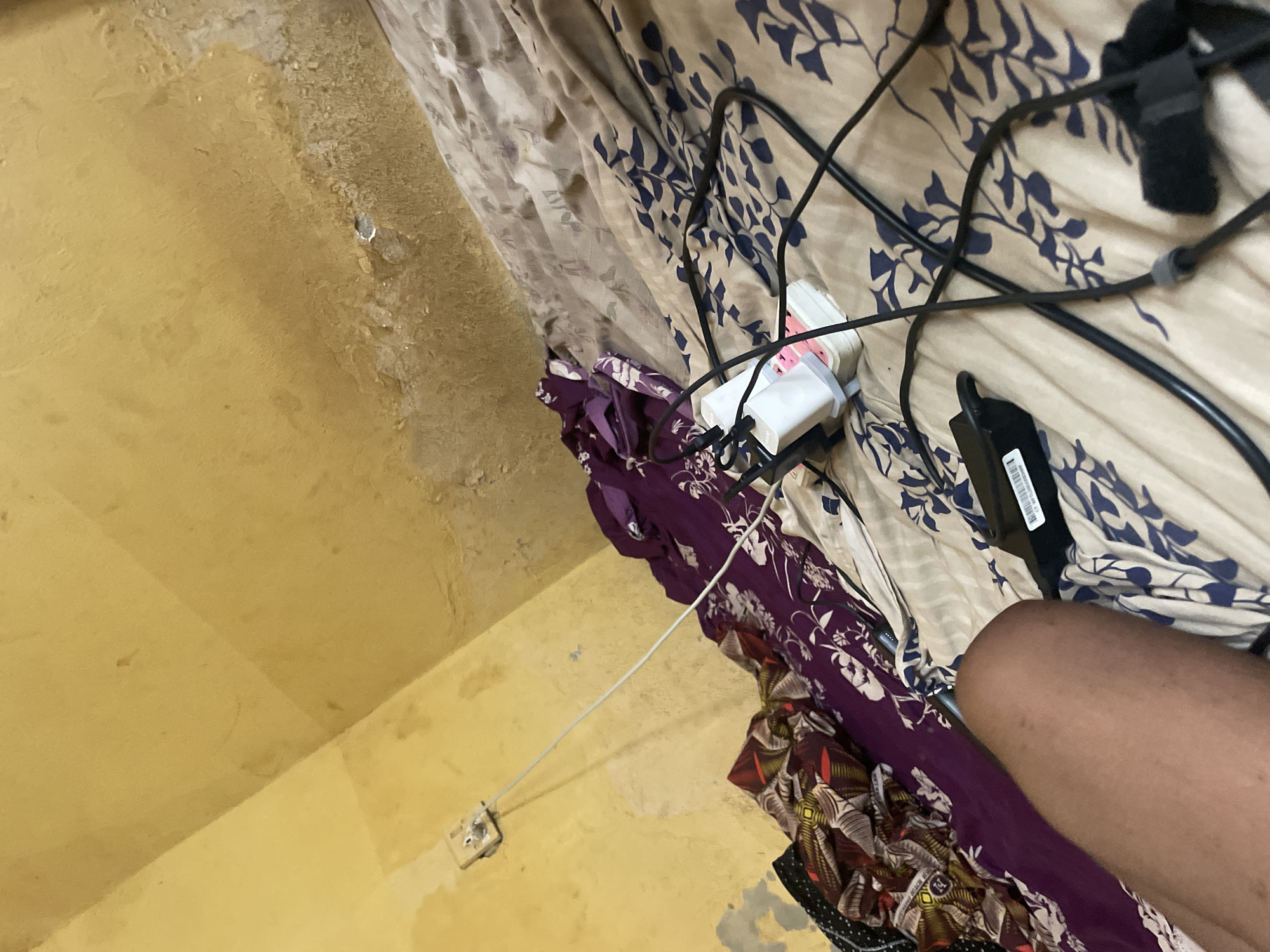 The image depicts a bedroom scene with a yellow, possibly damp, wall. On the wall, a power outlet is visible with a white cable extending from it. The bed is covered with patterned sheets, featuring a mix of floral and geometric designs in blue, beige, and maroon. Several items are strewn across the bed, including a purple garment, a power strip with multiple plugs, and various black cables. A person's leg, partially visible in the lower left of the frame, suggests someone is lying on the bed. The overall impression is one of a lived-in, potentially cluttered, domestic space. The location is Lokoja, Nigeria.