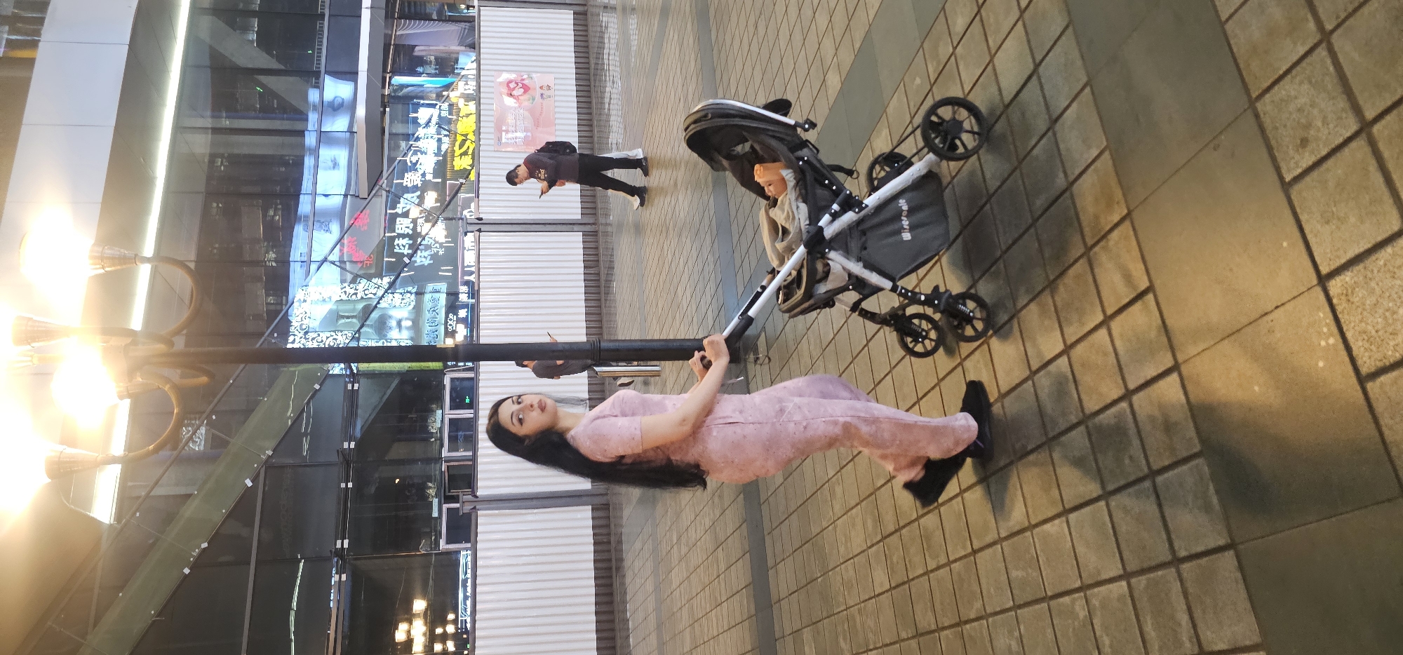 The photograph shows a woman pushing a stroller along a brick-paved sidewalk in what appears to be an urban area, likely Yuzhong District, China. The woman has long, black hair and is wearing a pink outfit. She is holding onto the stroller's handle, which appears to be attached to a child seat. Inside the stroller, there is an orange-colored object which appears to be a doll or a toy. Behind the woman and the stroller, a man is walking in the same direction, looking down at a mobile device in his hand. The background consists of modern buildings with illuminated signs, and architectural lighting.