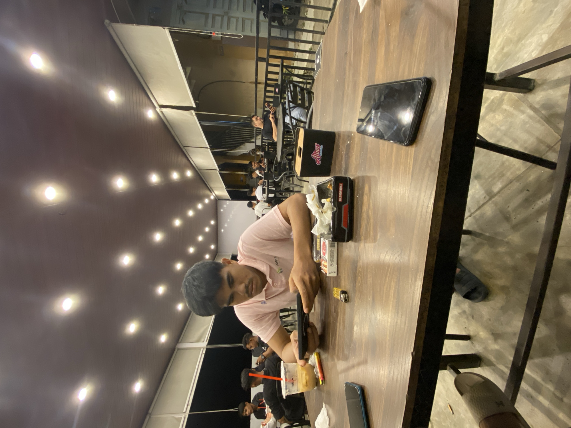 The image captures a casual nighttime scene in what appears to be an indoor or semi-outdoor cafe or restaurant in Keudeublang, Indonesia. The photograph is taken from a slightly elevated perspective, looking down at a wooden table and its occupants, and is rotated on its side.
In the foreground, a young man with dark hair, wearing a light pink polo shirt, is seated at a dark wooden table. He is intensely focused on a black smartphone he holds with both hands. To his right, on the table, is a small yellow lighter. Closer to him is a black box with "Aura" written in white cursive, and another black box with "BOLD NEW WORLD" visible on its side, along with the partially visible word "BATT". A plastic cup with a drink and a red straw is positioned further down the table to his left, next to another lighter and a phone.
The setting is illuminated by numerous circular white lights embedded in a dark brown or reddish-brown ceiling, creating a warm glow. In the background, other individuals, mostly men, are visible seated at tables, some also looking at their phones or socializing. A black metal railing separates the interior space from an exterior area where parts of a building and a motorcycle are faintly visible. The floor is concrete, and parts of chair legs are seen beneath the table.
The main activity is people relaxing and engaging with their mobile devices in an evening social setting. The presence of food packaging and drinks suggests a dining or refreshment establishment.
