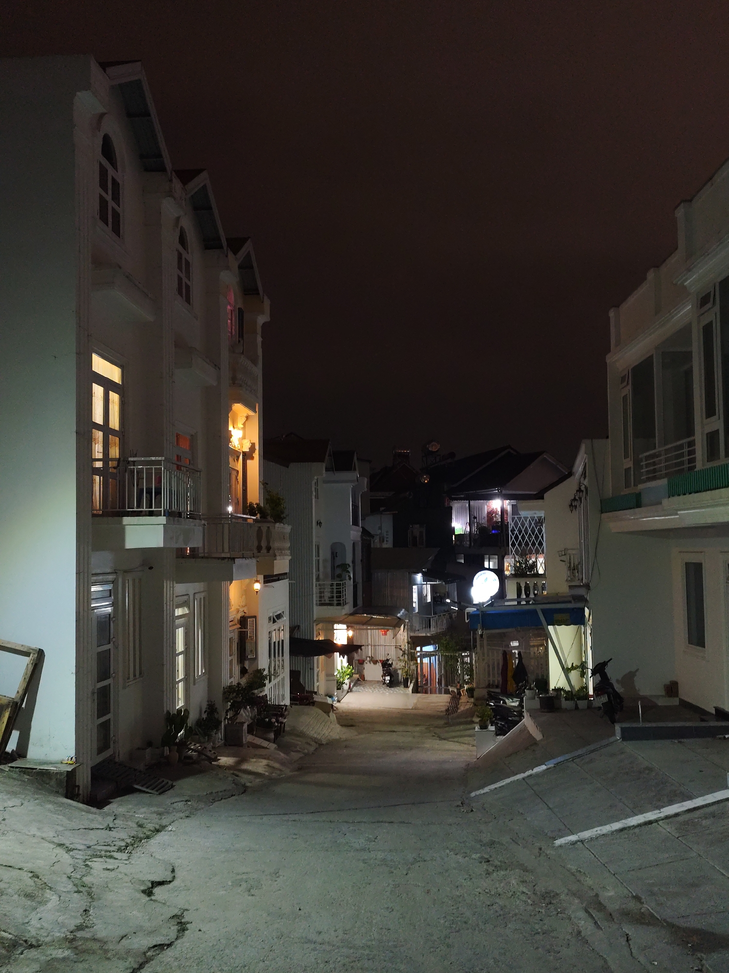 Captured at night, the photograph reveals a street in Da Lat, Vietnam, flanked by multi-story buildings. The buildings, primarily white, display architectural details such as arched windows and balconies. Illumination comes from internal lights within the buildings, as well as streetlights and lights from businesses further down the street. The street itself is paved and shows signs of wear and cracking. Several parked motorcycles are visible along the street. The sky is dark, suggesting a late hour. Vegetation, including potted plants, is placed along the street and in front of buildings.
