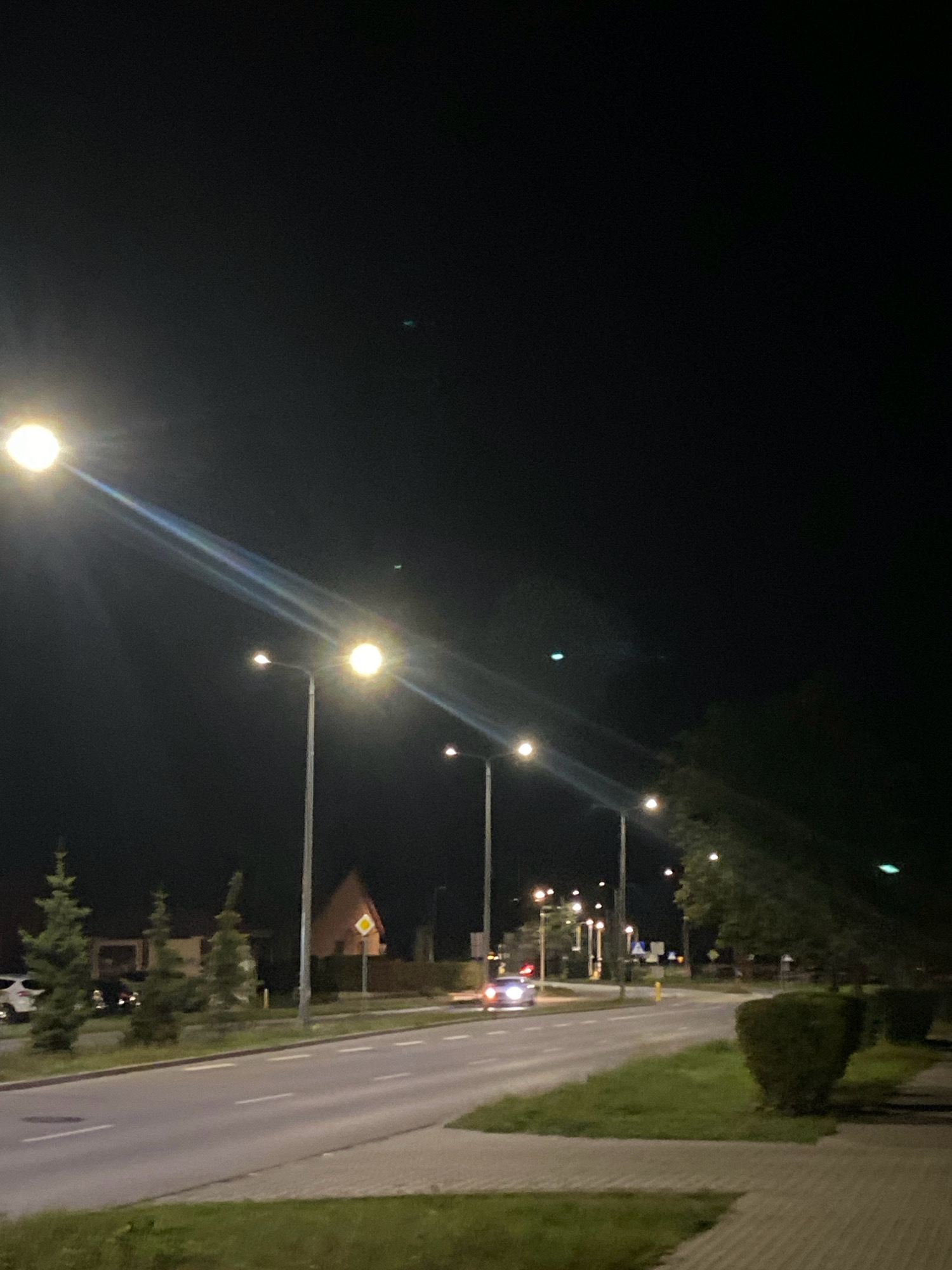 The image captures a nighttime street scene with multiple streetlights illuminating the road and surrounding area. In the foreground, a paved road runs diagonally from the lower left to the upper right. To the left of the road, there is a grassy area with several coniferous trees and parked cars. Across the road, on the right side, is a sidewalk and more grassy areas with bushes. Further down the road, on the left, a building with a triangular roof and a diamond-shaped sign is visible, suggesting it might be a residential or commercial area in Nowy Sącz, Poland. A vehicle with its headlights on is driving on the road, and several other streetlights line the street at intervals. The sky is dark and clear, with no visible stars or moon. The overall impression is of a quiet, well-lit urban environment at night.