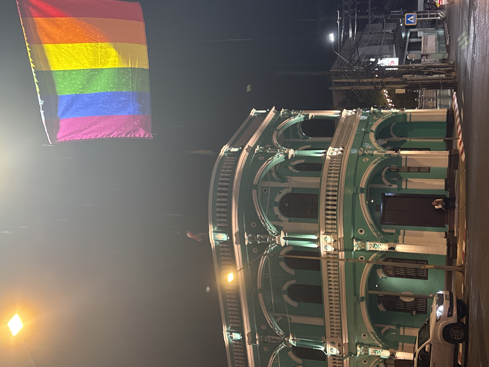 A nighttime scene in Phuket City, Thailand, is captured in this image, likely on a street in the Old Town given the architecture. The main subjects are a vibrant rainbow flag, a striking green colonial-style building with white accents, and a white pickup truck. The flag, a symbol of LGBTQ+ pride, hangs prominently in the upper left of the frame.
The building itself is a key element, showcasing ornate balconies, arched windows, and decorative columns, illuminated by warm lights, suggesting it might be a shop, restaurant, or residential space. A single person is visible near the entrance of the building, possibly a resident or patron. The white pickup truck is parked on the right side of the road, its headlights on.
The setting appears to be a city street at night. The sky is dark, and artificial lights from streetlamps and the building illuminate the scene. The street surface appears wet, possibly indicating recent rain or a humid climate. Overhead, a complex network of electrical wires and streetlights is visible, typical of many Southeast Asian cities. A blue and white pedestrian crossing sign is also present in the background, further contextualizing the urban setting.
There is no discernible text in the image, and the overall mood is calm and perhaps a bit mysterious due to the nighttime setting and lighting. The focus is on the architectural beauty of the building and the symbolic presence of the rainbow flag, suggesting a sense of inclusivity or celebration in this locale.