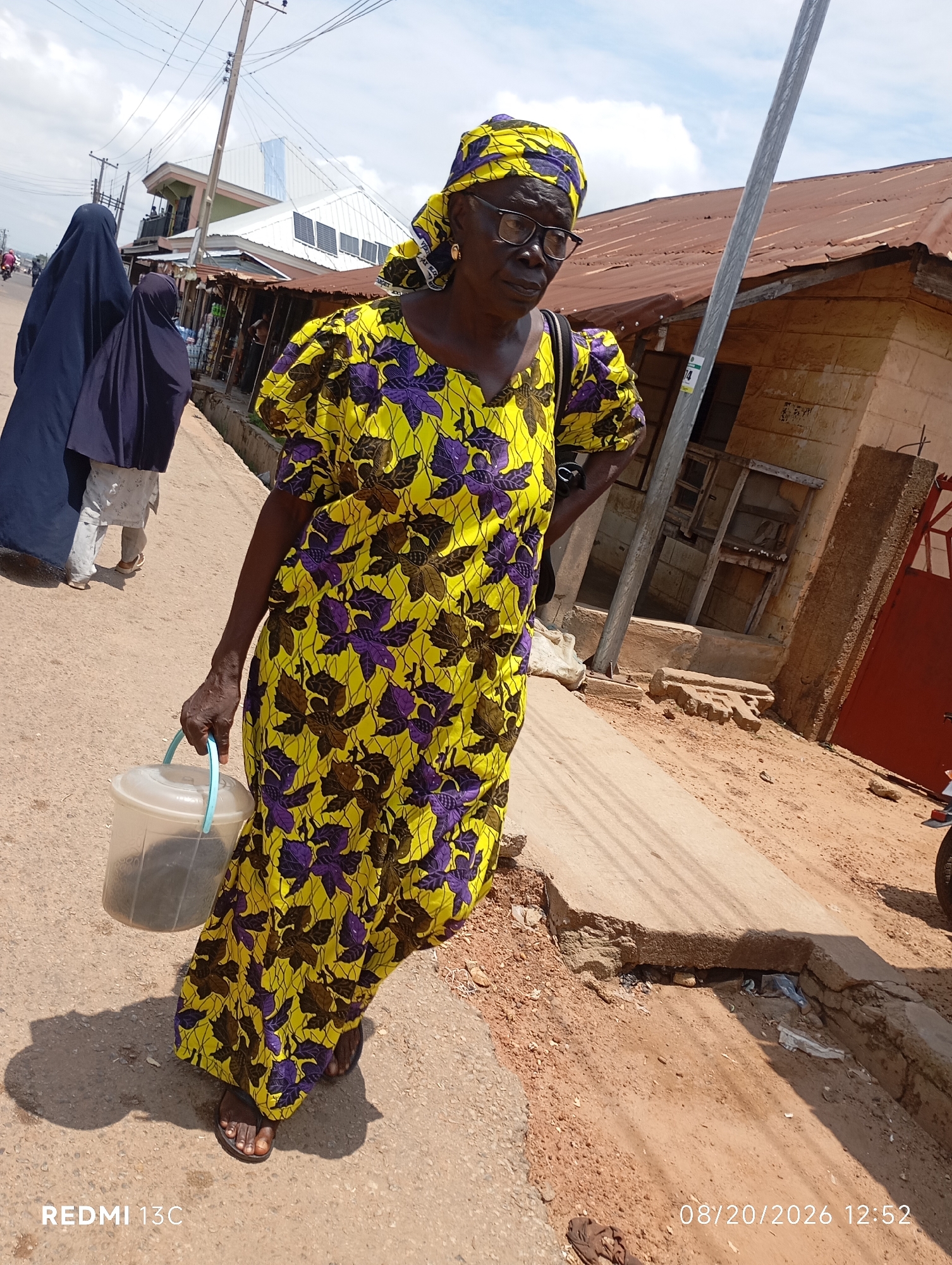 In the city of Shendam, Nigeria, under a bright sky with scattered clouds, a Black woman in her late 60s, wearing a vibrant yellow and purple floral dress and a matching headscarf, walks along a dusty, uneven street. She is holding a transparent plastic bucket with a light blue handle in her right hand, its contents not clearly visible. Her feet are clad in dark sandals. Behind her, two individuals wearing dark blue hijabs walk further down the street, their faces obscured. The environment features a mix of buildings, one with a corrugated iron roof and another with a modern white facade, suggesting a developing urban area. There are utility poles and wires overhead, and the ground is a mixture of packed dirt and cracked concrete. The overall scene captures a moment of daily life in a Nigerian town, with evidence of both traditional elements and ongoing development.