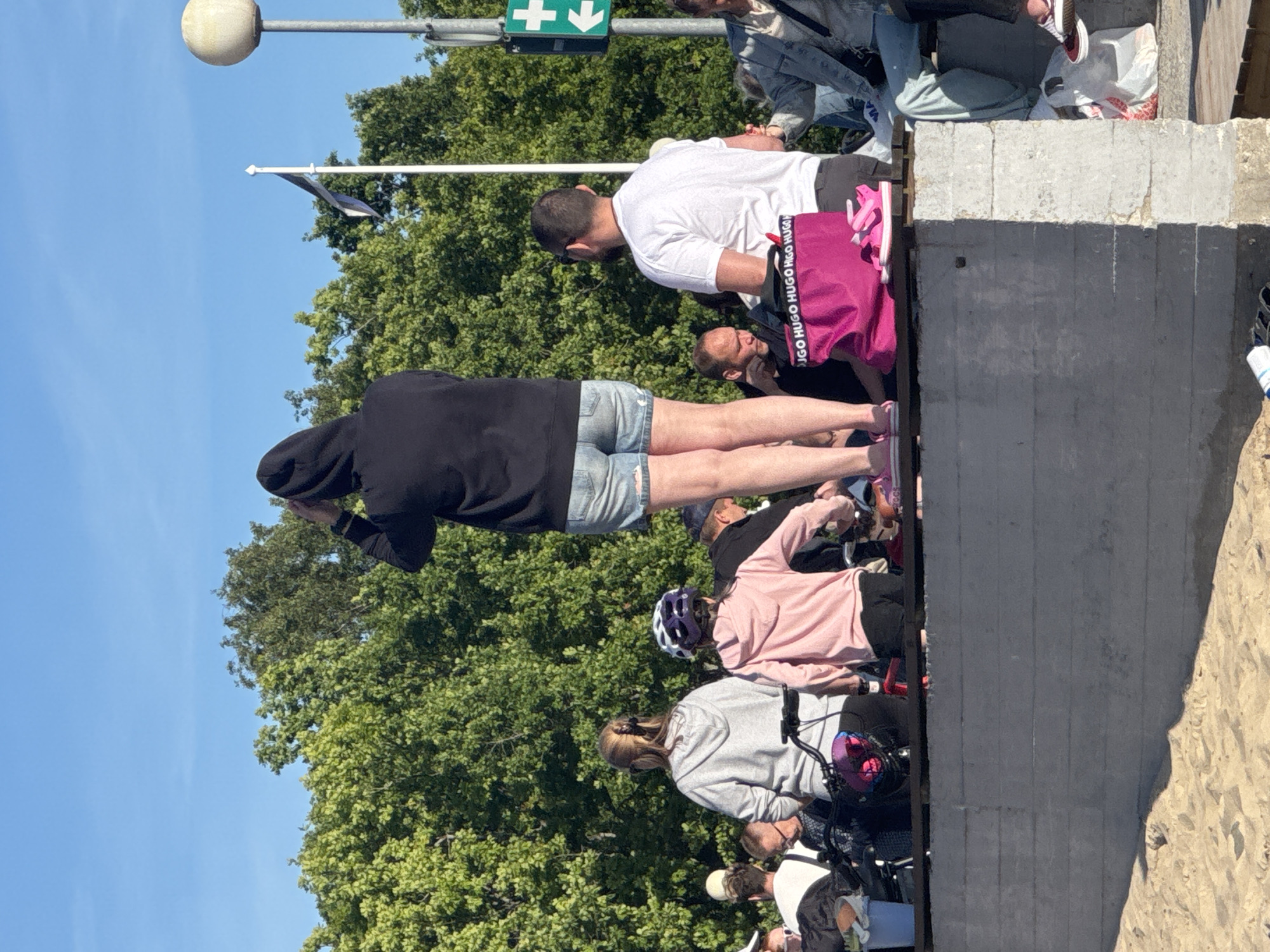 This image depicts a group of people gathered on a raised concrete structure, possibly a promenade or a seating area, near a beach with sand in the foreground. The background features lush green trees and a bright blue sky. In the mid-ground, several individuals are visible, including a person in a black hoodie and denim shorts looking upwards, a woman in a grey hoodie, a child in a pink hoodie and helmet, and a man in a white t-shirt. Other individuals are partially visible, some seated and others standing. A bright pink bag with "HUGO HUGO HUGO" text is prominently displayed. A flagpole with a flag, a lamppost with a spherical light, and a green cross sign with an arrow are also present in the scene. The overall atmosphere suggests a casual gathering on a sunny day, likely in an outdoor public space in Tallinn, Estonia, given the context of the surrounding environment and potential local landmarks.