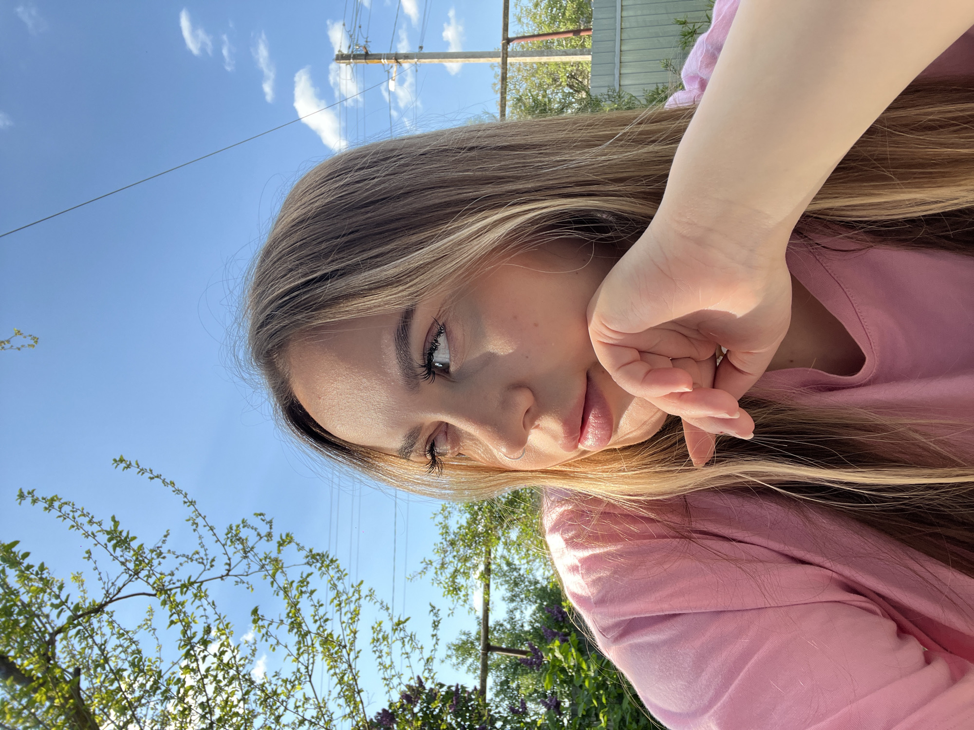 A woman is positioned in a slightly elevated outdoor location in Novopokrovskaya, Russia. She is fair-skinned and has long, blonde hair. Her head is tilted, and she rests her hand on her face, looking downward with one eye visible. She wears a pink long-sleeved shirt. The sky is blue with some white clouds. Below the woman, greenery and tree branches are visible. A utility pole with wires stretches across the upper portion of the frame. A metal fence or structure appears in the background.