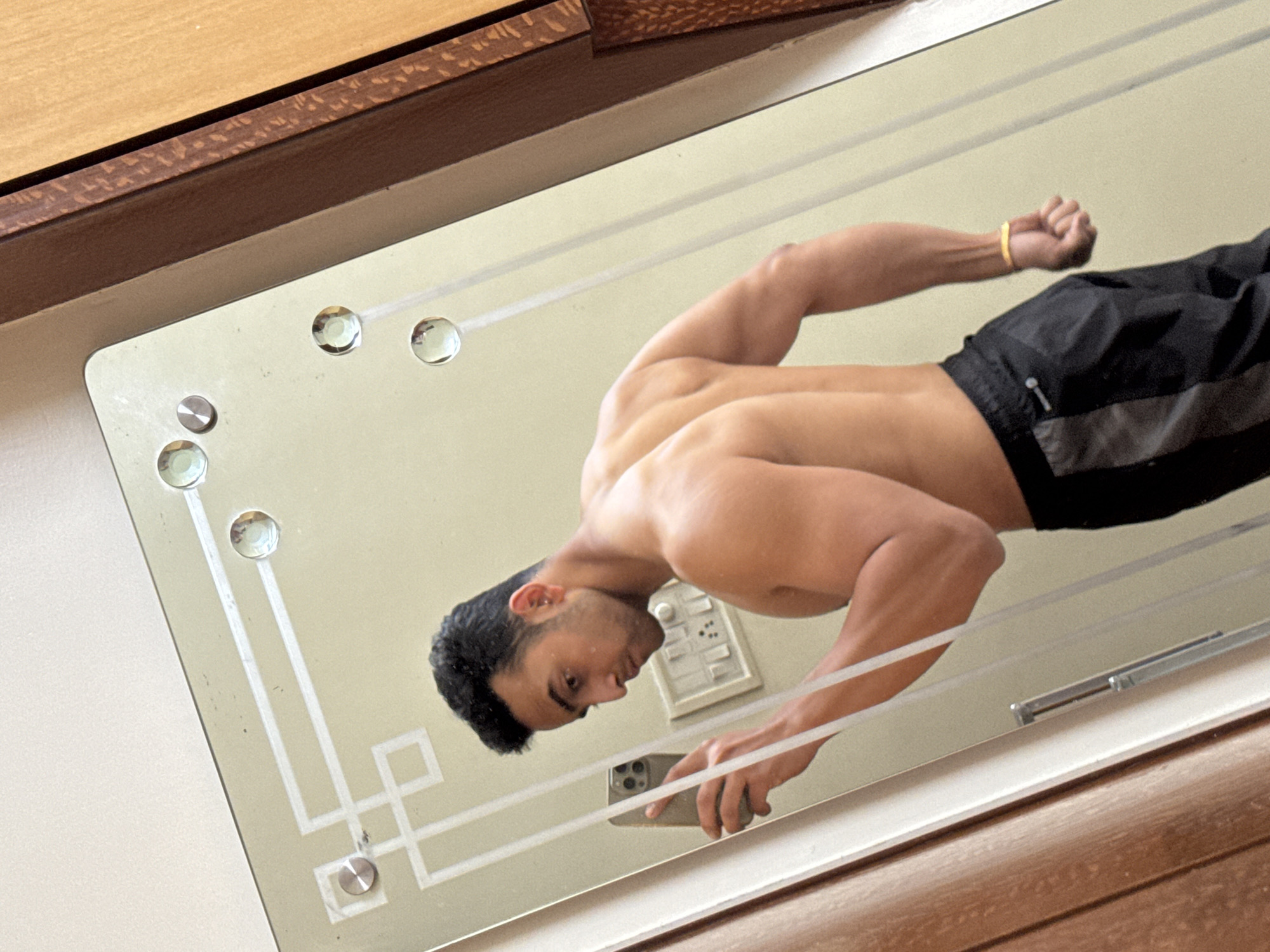 The image displays a shirtless, muscular male subject taking a mirror selfie inside an unidentified interior space in Kolkata, India. The subject is positioned with his back and left side towards the viewer, showcasing his developed back and arm musculature. He is wearing black athletic shorts with grey panels. His right hand holds a smartphone, which is angled to capture his reflection. A thin, gold-colored bracelet is visible on his left wrist. The mirror features an etched geometric pattern in the upper left quadrant and is secured by multiple circular, presumably metallic, fasteners. Reflected in the mirror, behind the subject's left shoulder, is a white electrical switchboard containing several switches and power outlets, mounted on a light-colored wall. Wooden panels or furniture are visible flanking the mirror's reflection on both the left and right sides. No specific external landmarks or architectural details are visible to pinpoint a precise location within Kolkata.