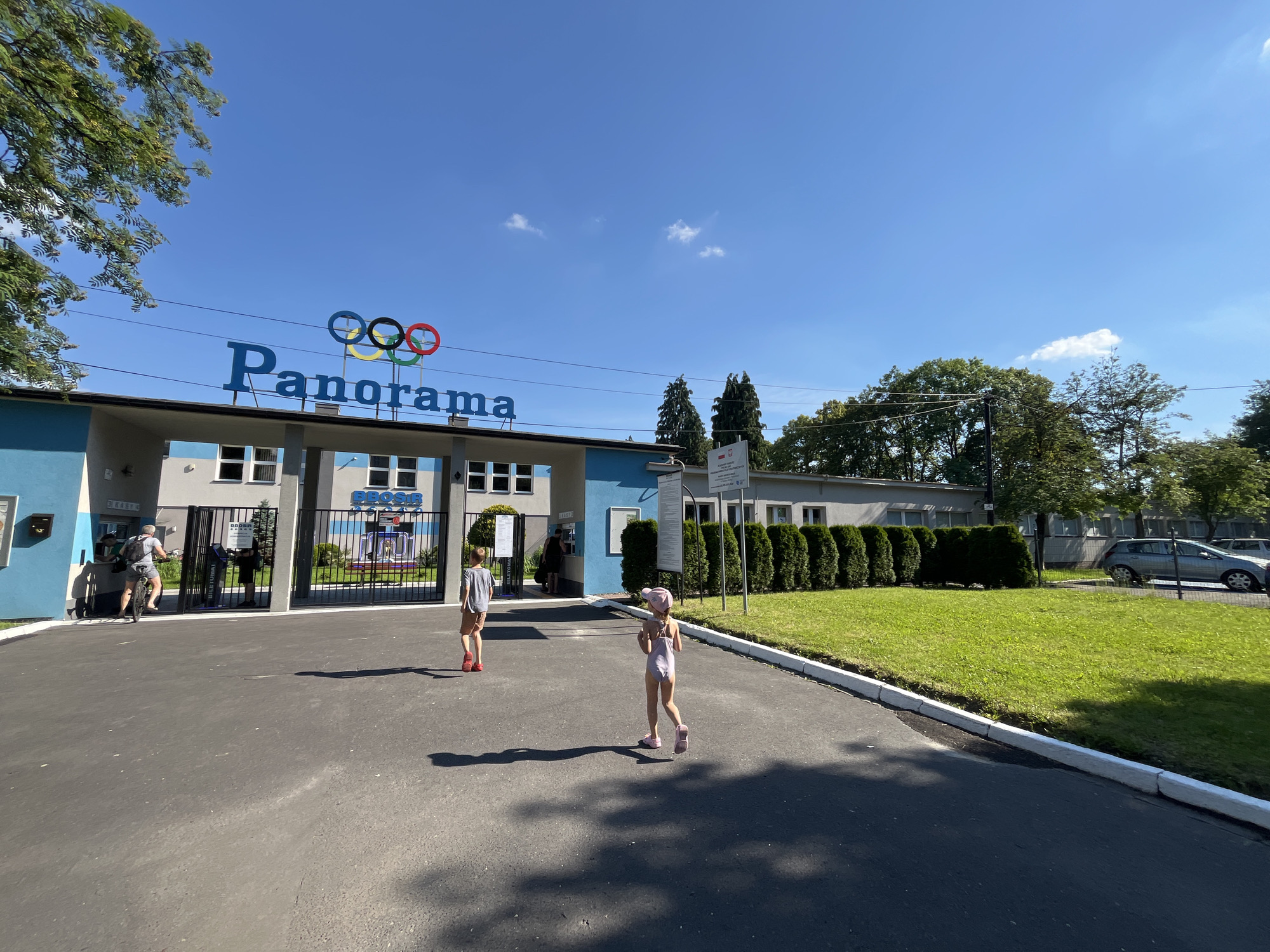 A low-angle, wide shot captures the entrance of a facility labeled "Panorama" under a clear blue sky. The "Panorama" text is in large blue letters, with the Olympic rings positioned above and to the left of the text. The building entrance is painted in white and blue with multiple glass-paned windows. A black metal gate is open, with people visible beyond it. In the foreground, a paved pathway leads towards the entrance. Two children are walking away from the camera on the pathway; one is wearing a light-colored swimsuit and a pink hat, while the other is wearing a grey t-shirt and shorts. To the right of the pathway is a well-maintained green lawn bordered by a white curb. In the background, a line of evergreen bushes separates the lawn from a building with a light-colored facade and more windows. A silver car is parked in the background, to the right of the evergreen bushes. The environment appears to be an outdoor recreational area or a public facility on a sunny day.