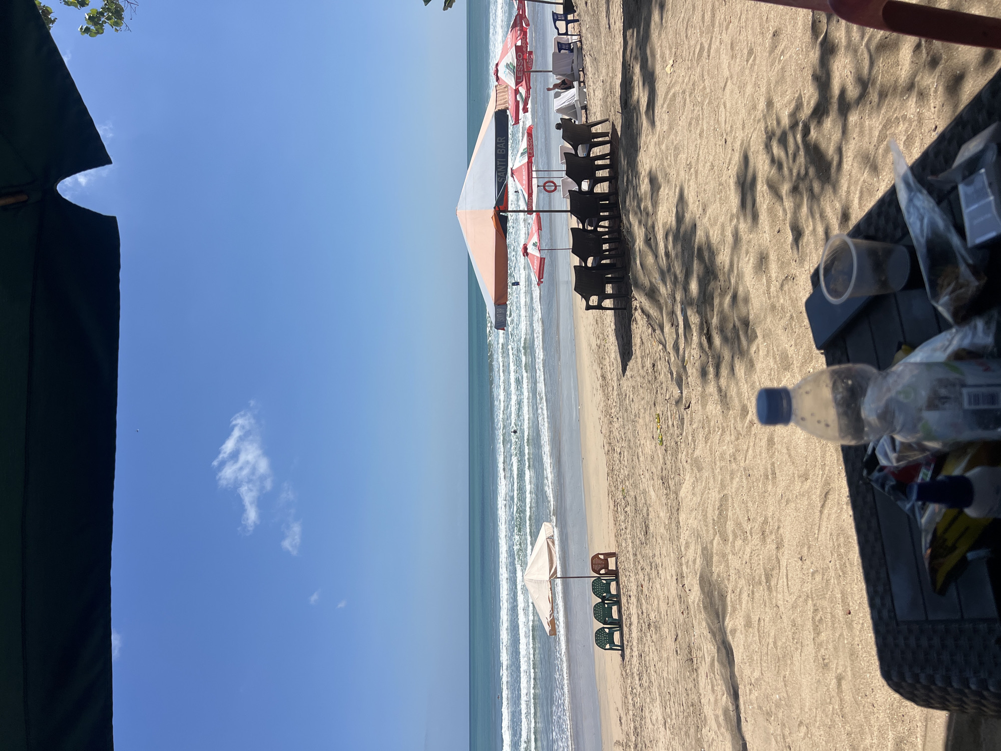 The image captures a sunny day at Kuta Beach, Indonesia, with the foreground occupied by a table set up on the sand. On the table, a partially filled plastic water bottle, a clear plastic cup, a smartphone, and various bagged items are visible, suggesting a relaxed beachside setting. The sandy beach stretches towards the ocean, where gentle waves are breaking. In the mid-ground, several rows of beach chairs and umbrellas are arranged. One set of chairs, consisting of three green plastic chairs and one brown plastic chair, is positioned under a light-colored umbrella closer to the water. Further back, a larger, multi-colored umbrella is set up, with more chairs and lounge chairs visible underneath and around it, some occupied by individuals. The sky is clear blue with a few scattered white clouds, and the ocean appears calm with a few surfers in the distance. The overall impression is one of a typical tourist beach scene in Kuta, Bali.