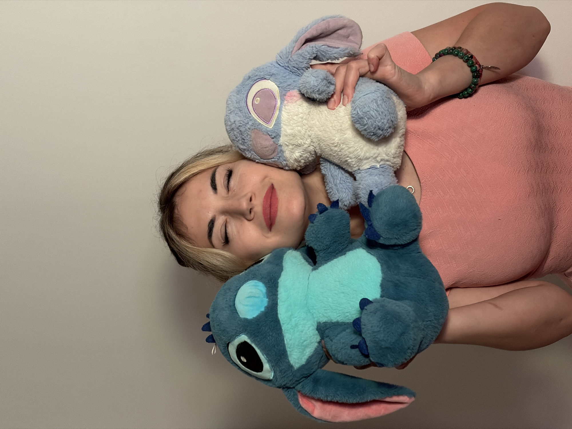 A young woman with blonde hair, dressed in a pink top, is pictured against a plain, light-colored wall. She is holding two plush toys: one light blue and white resembling Angel, and another darker blue and light blue resembling Stitch, both from the Lilo & Stitch franchise. Her eyes are closed, and a soft smile graces her face, conveying a feeling of contentment or affection. A beaded bracelet, featuring green and reddish-pink beads, adorns her right wrist. The scene suggests an indoor setting, likely in Budva, Montenegro, with soft, ambient lighting. No text is visible in the image.