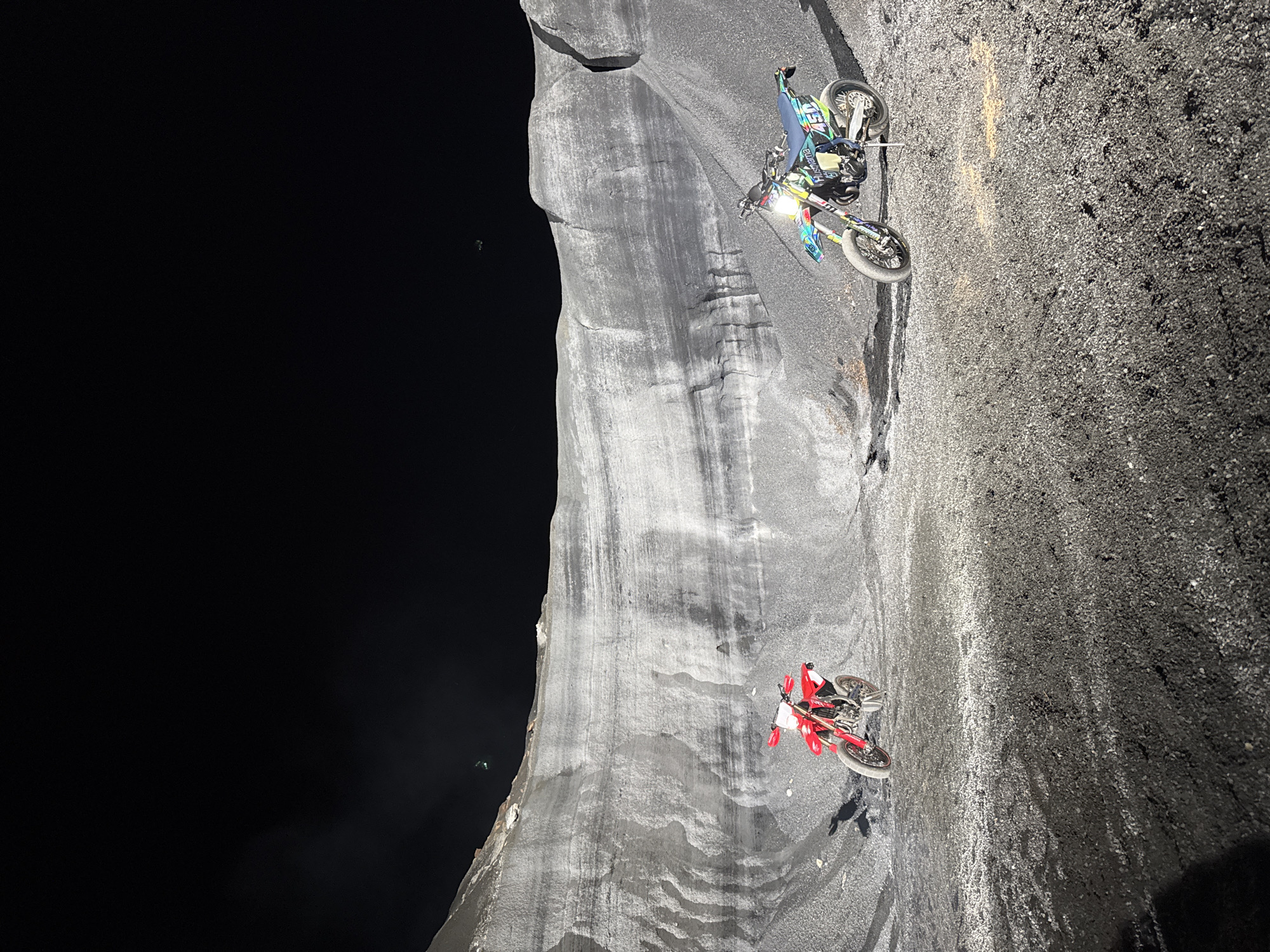 Two dirt bikes are parked on a gray gravel surface. The surface seems to form the edge of a cliff. To the left of the cliff edge is a dark, shadowy void. The top bike is predominantly blue and green, while the lower one is red. The setting appears to be outdoors, possibly near a quarry or a natural rock formation in Teguise, Spain. The people cannot be seen clearly.