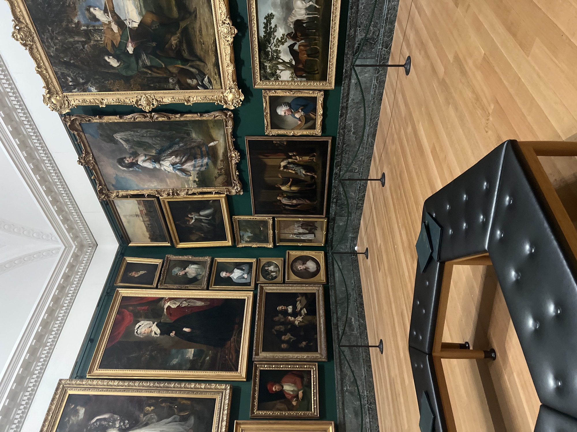 The image displays an interior view of an art gallery or museum, with a slightly elevated perspective looking into the room. The walls are painted a dark green, serving as a backdrop for a densely hung collection of paintings in a salon-style arrangement. The floor is made of polished light-toned wooden planks. White decorative ceiling molding with intricate patterns is visible along the top left edge.
The main subjects are numerous framed oil paintings of various sizes. Many are portraits, depicting individuals such as a man in a blue coat holding a circular object, women in historical attire (e.g., one in a blue and white striped dress, another in a black dress with a white cap), and other figures. Several paintings also portray genre scenes with multiple people engaged in activities, including a large piece depicting several figures in what appears to be a historical or religious context. A few landscape paintings, such as one featuring horses, are also present. All artworks are enclosed in elaborate, gilded frames.
In the lower right, a dark, possibly leather-upholstered, multi-faceted bench or seating unit is visible, featuring wooden legs, characteristic of museum seating. Metal stanchion bases are intermittently placed on the wooden floor, likely indicating a viewing distance or forming a barrier. The scene depicts a quiet, formal art exhibition space. There is no visible activity, and no people are present in the frame. No discernible text is visible within the paintings or on the walls. The indoor setting is illuminated by artificial gallery lighting.